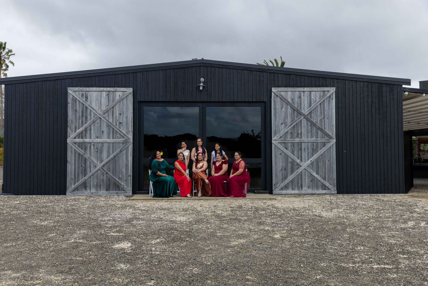 Bridal party in red dresses posing outside a dark barn venue with rustic wooden doors on a cloudy day.