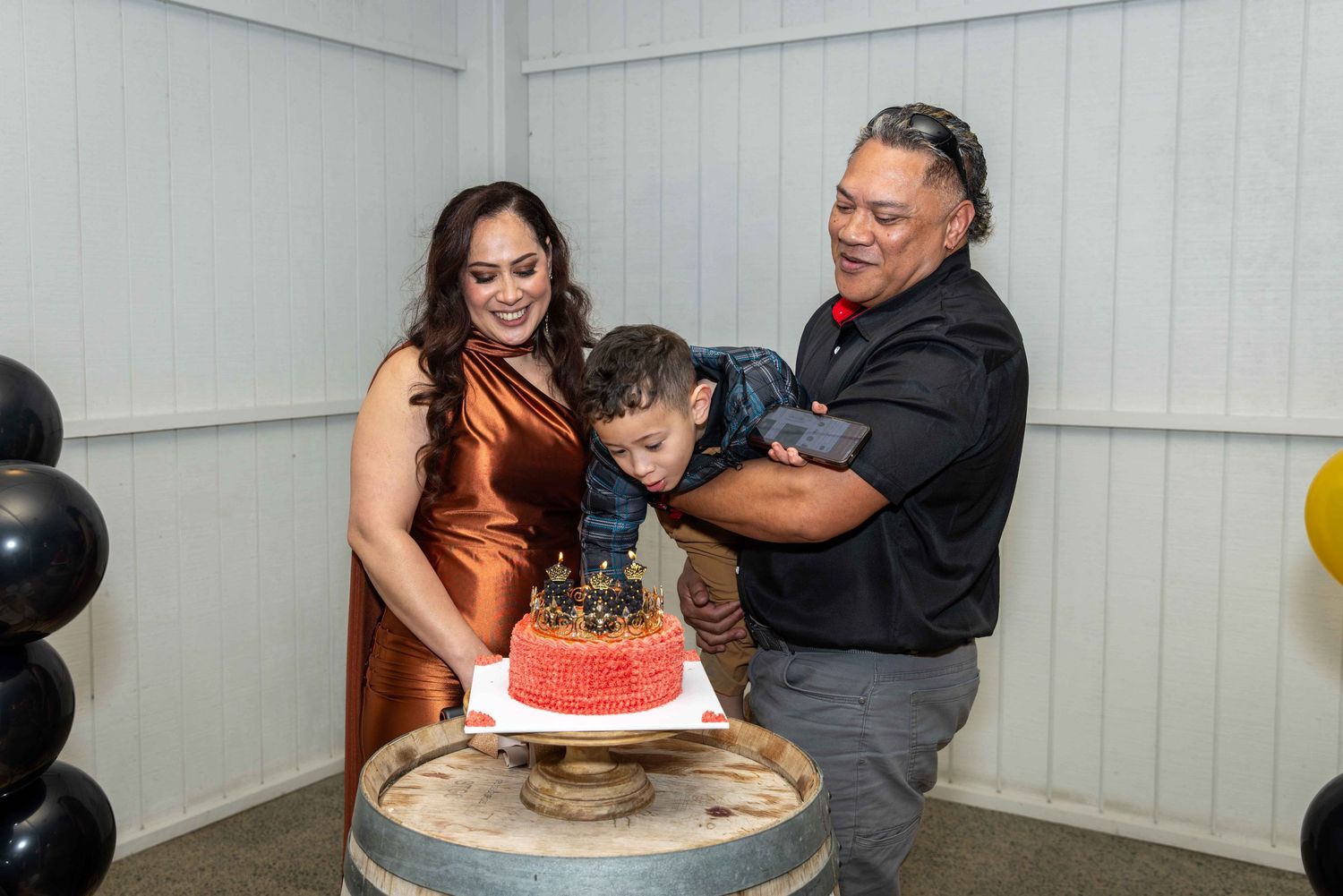Family celebrating a child's birthday with a red cake on a barrel, surrounded by black and gold balloons.