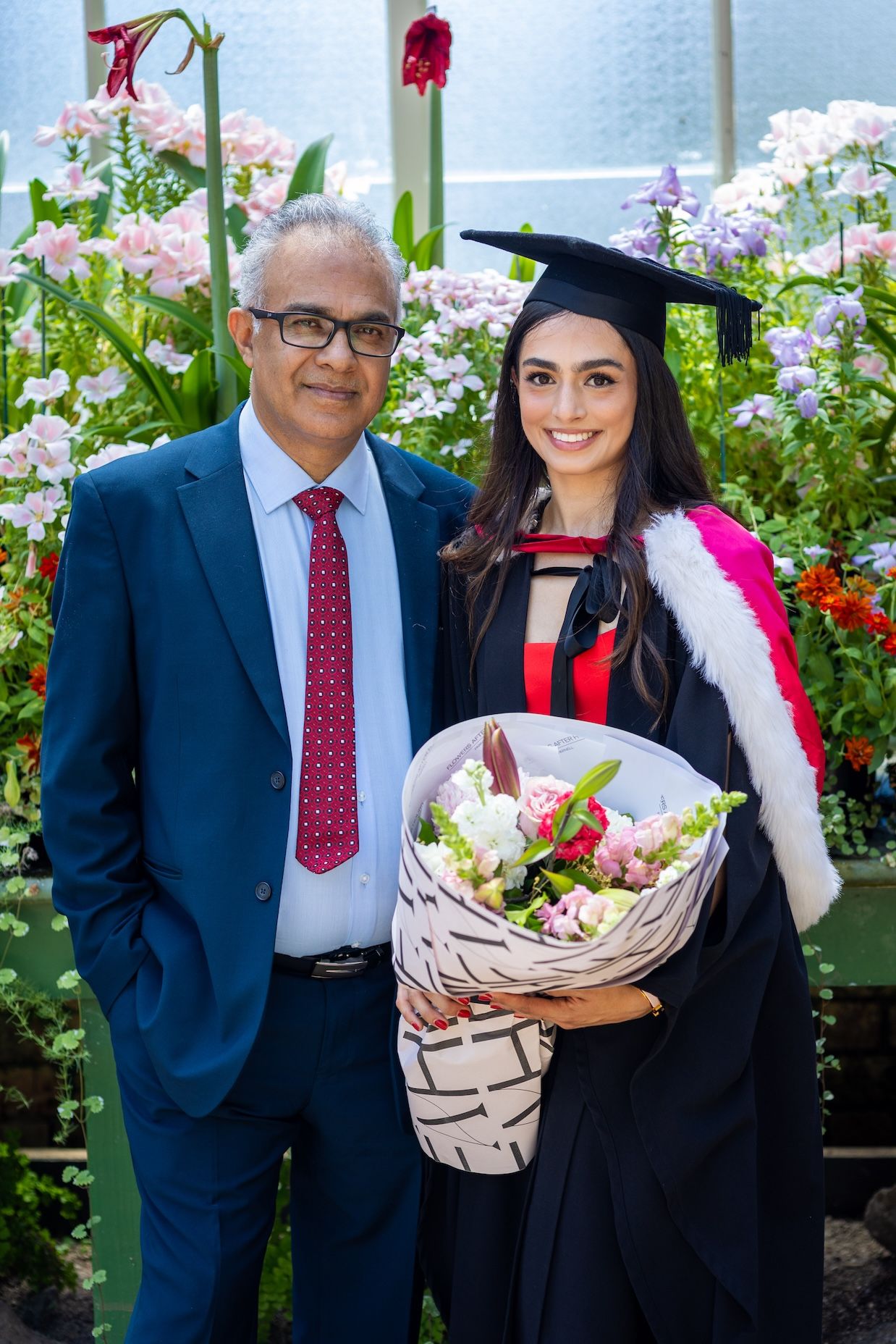 Graduate in cap and gown holds flowers beside older man in blue suit at graduation ceremony.