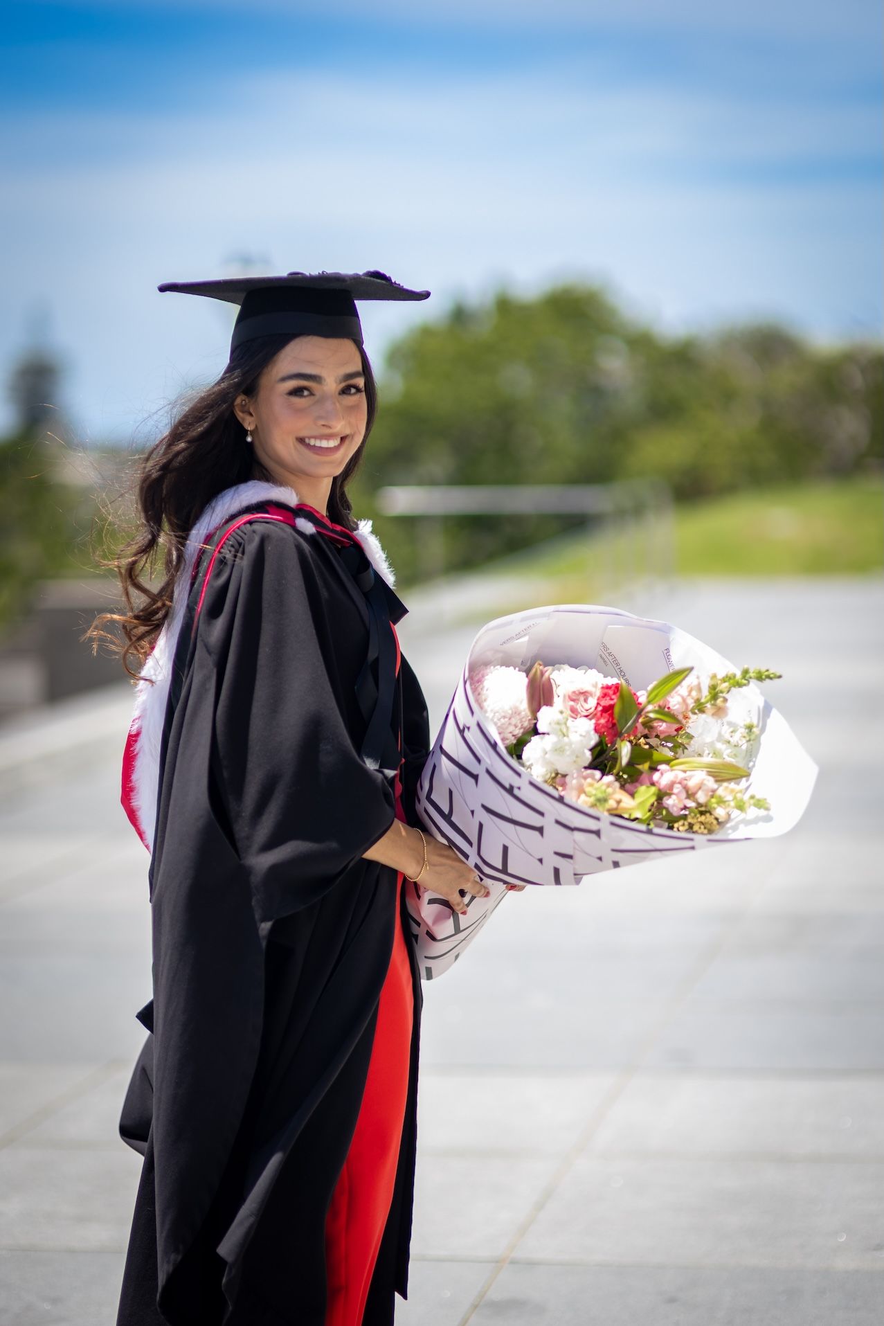 Smiling graduate in black gown and cap holds a bouquet of flowers outdoors on a sunny day.