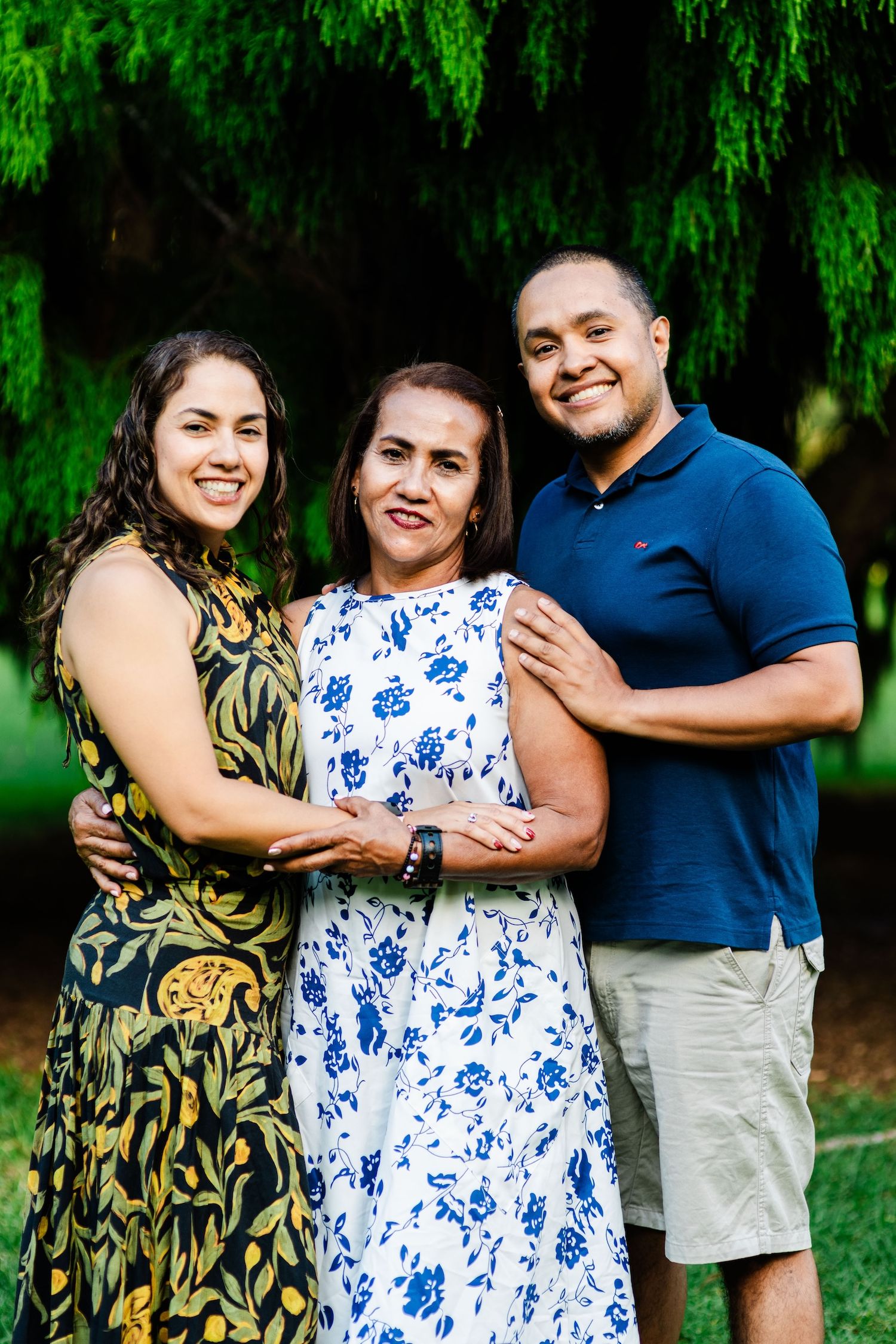 Three smiling family members pose together outdoors under lush green trees, wearing casual summer attire.