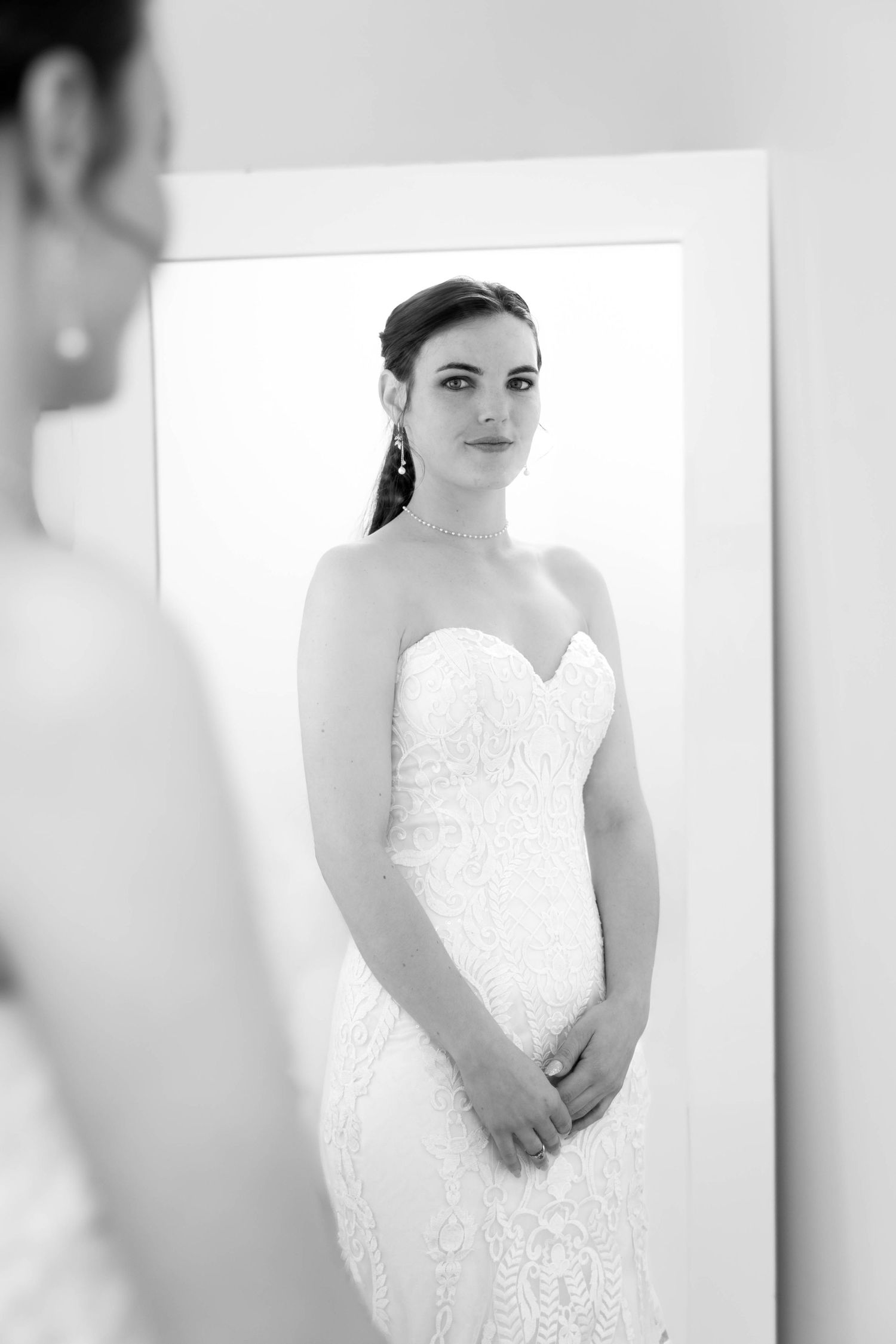 Bride in strapless lace wedding gown gazes at her reflection in a large mirror, black and white photo.