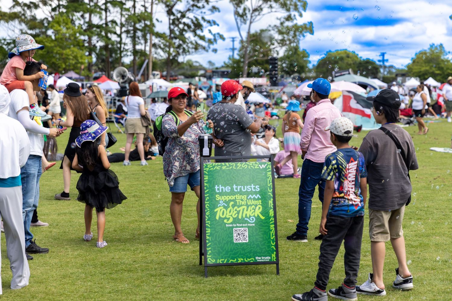 Large crowd gathers on a grassy field with green promotional banners at an outdoor event.