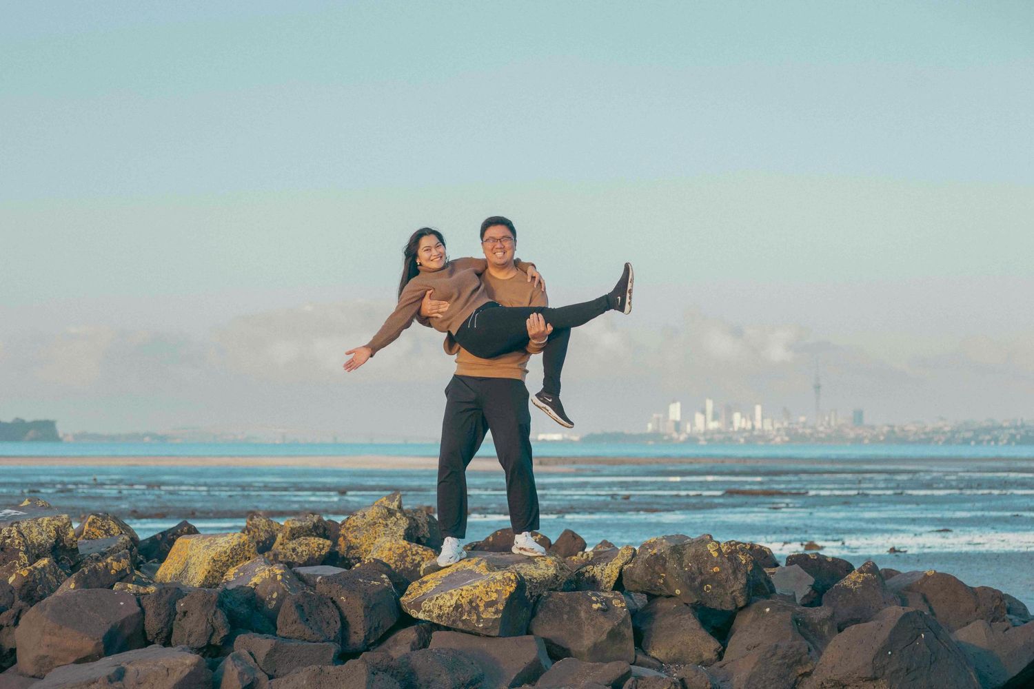 Couple posing on rocky shoreline, man carrying woman piggyback with ocean and city skyline backdrop.