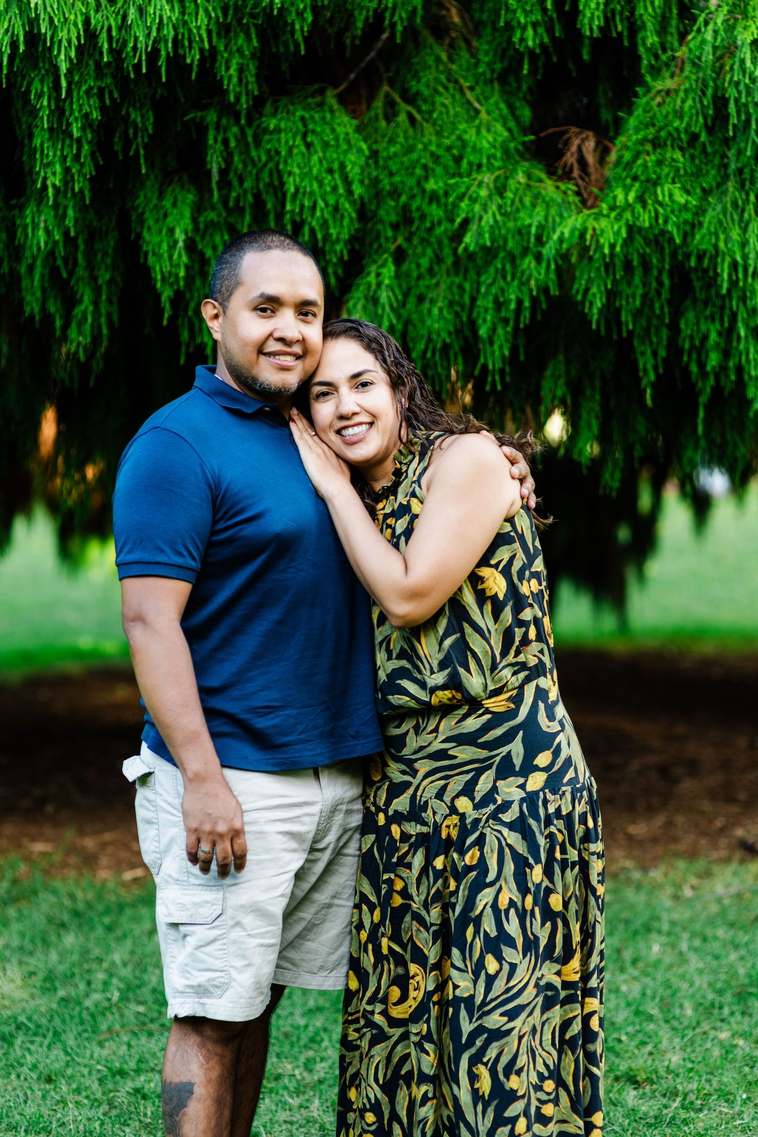 A smiling couple poses outdoors under a weeping tree, man in blue polo and khaki shorts, woman in floral dress.
