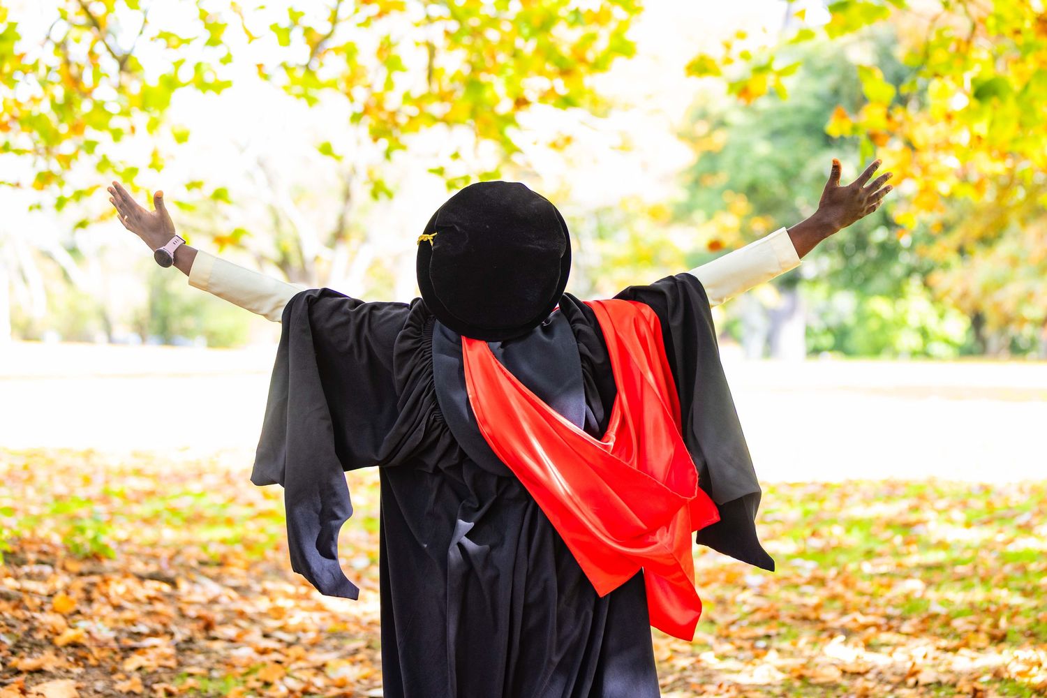 Graduate in black gown and red sash celebrating outdoors with arms outstretched among autumn foliage.