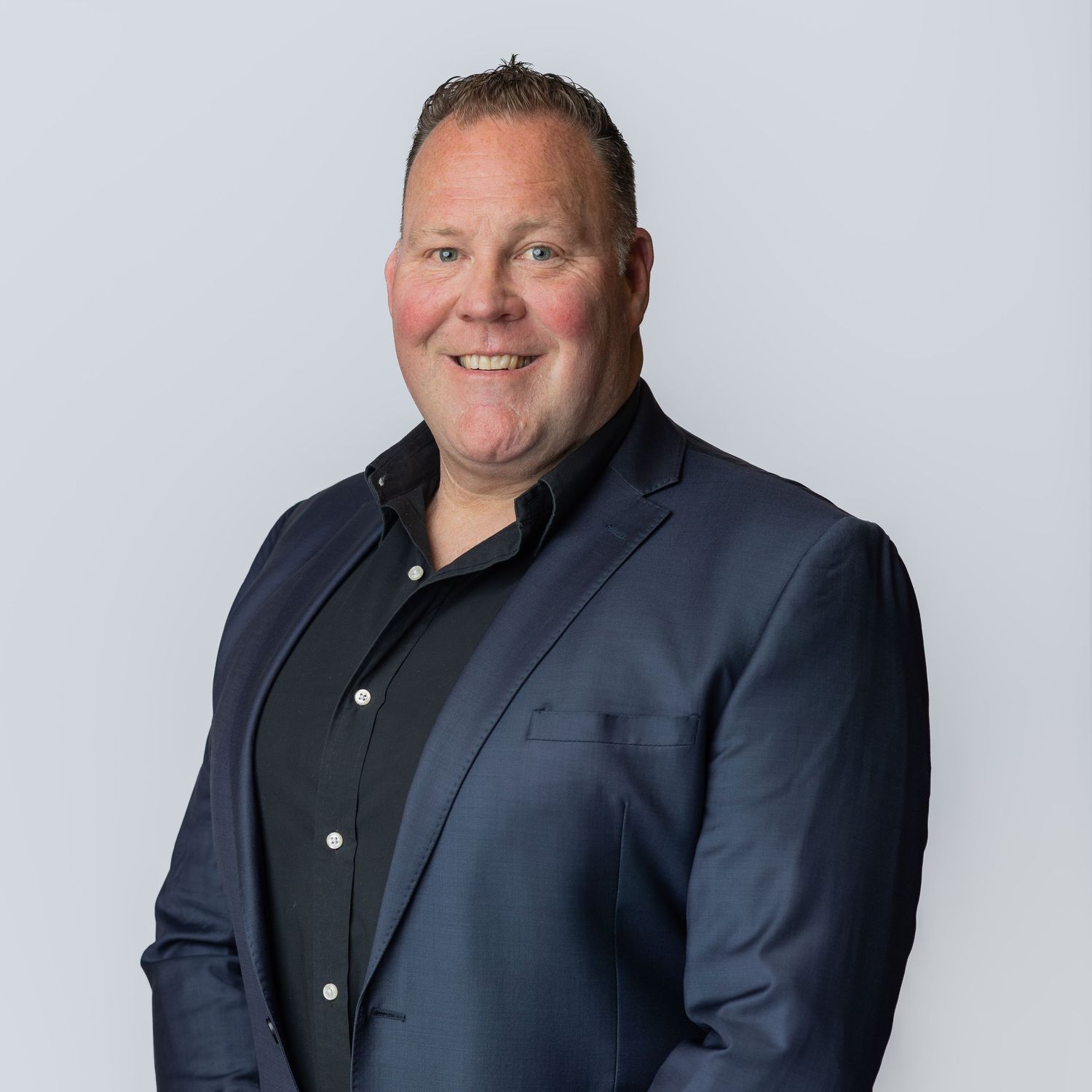 A smiling middle-aged man in a navy blue blazer and black shirt poses for a professional headshot against a gray background.
