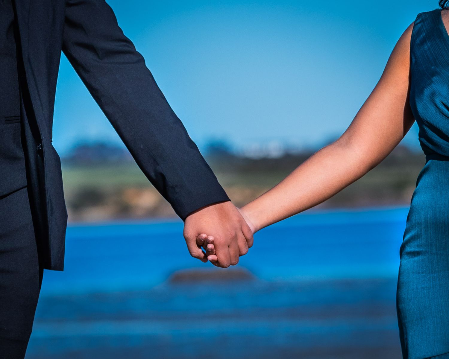 Couple holding hands outdoors near blue water, man in dark suit and woman in teal dress, romantic moment.
