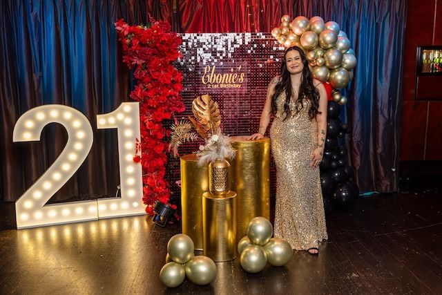 Woman in gold gown poses at elegant 21st birthday party with balloon arch, floral wall, and marquee number decorations.