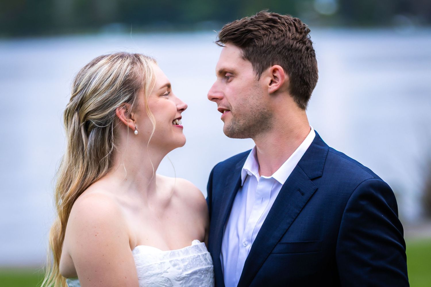A smiling bride and groom share a romantic moment outdoors near a lake, dressed in wedding attire.