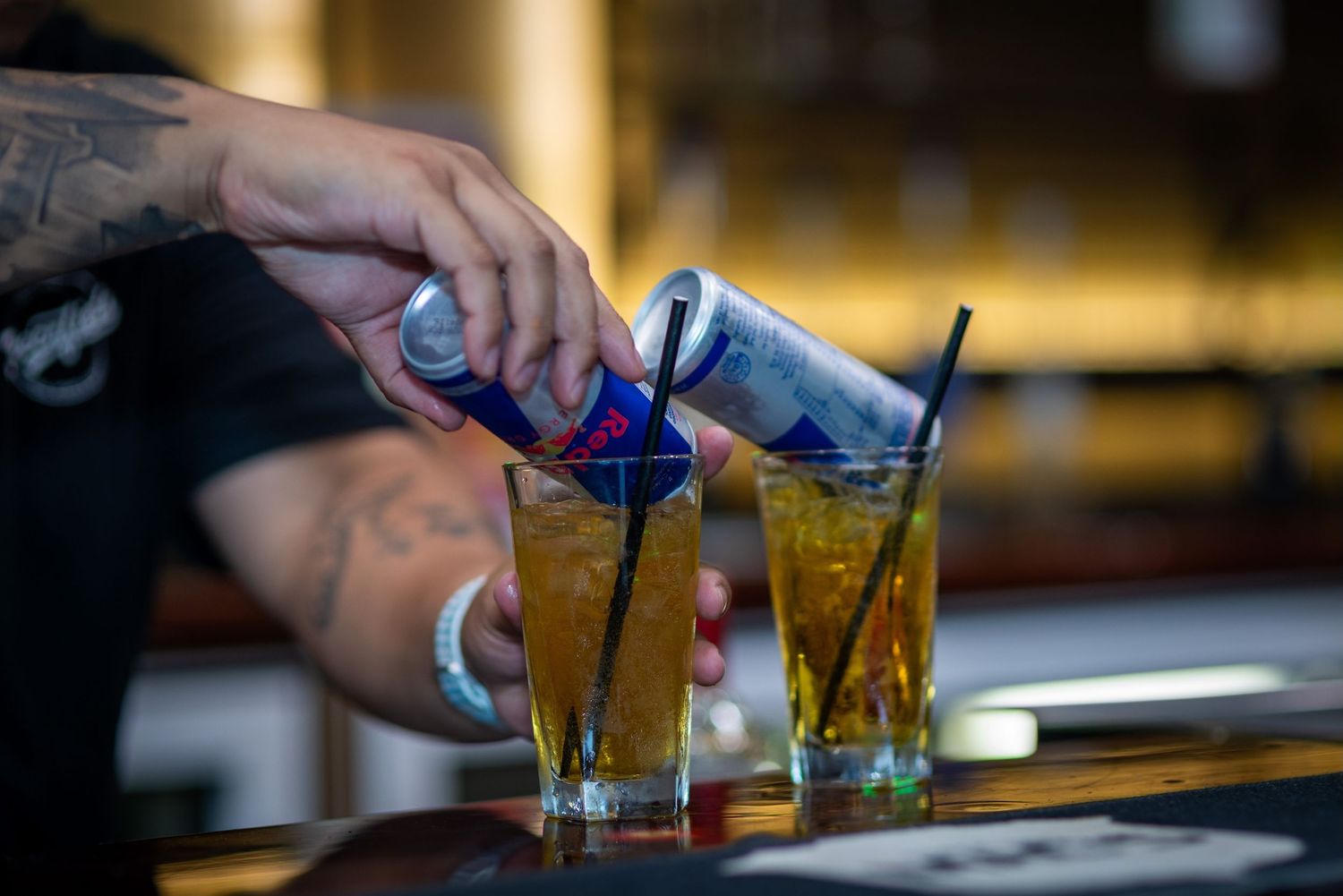 A bartender pours blue liqueur into two tall cocktail glasses filled with ice and amber-colored drinks.