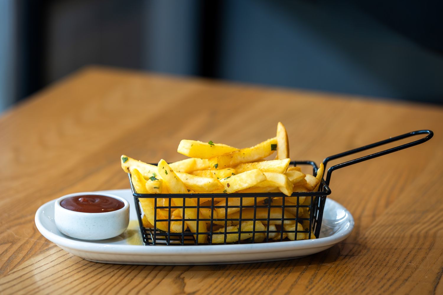 Golden french fries served in a white dish next to dipping sauces on a wooden table.