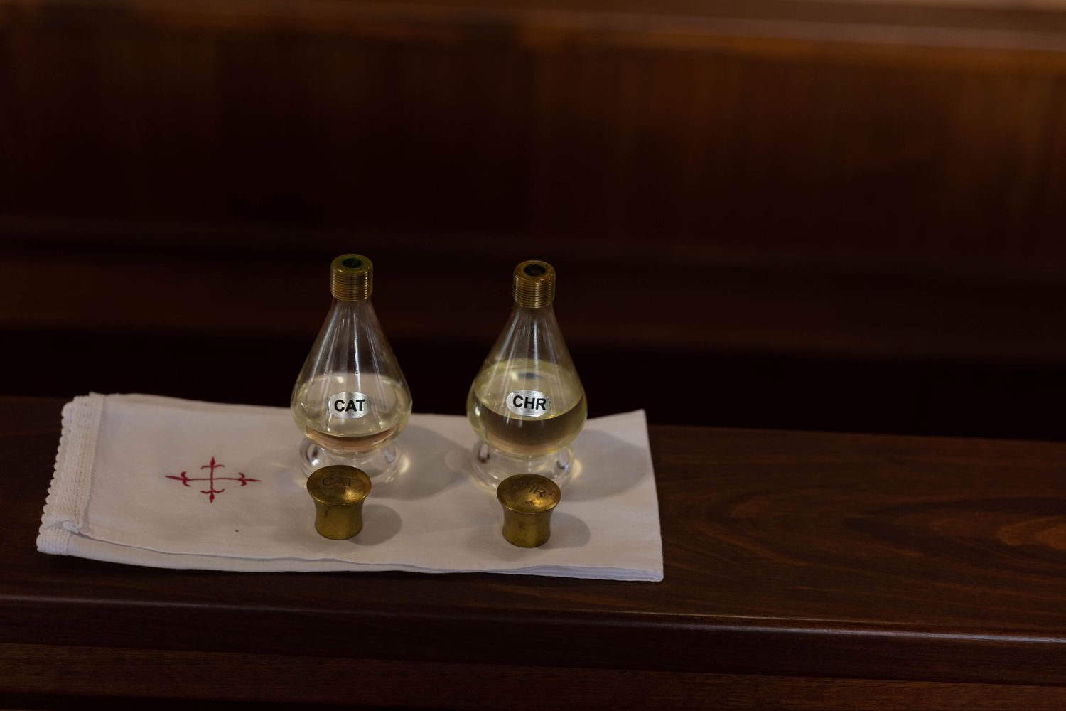 Two glass cruets with brass caps on a white liturgical cloth with red cross embroidery on a wooden surface.