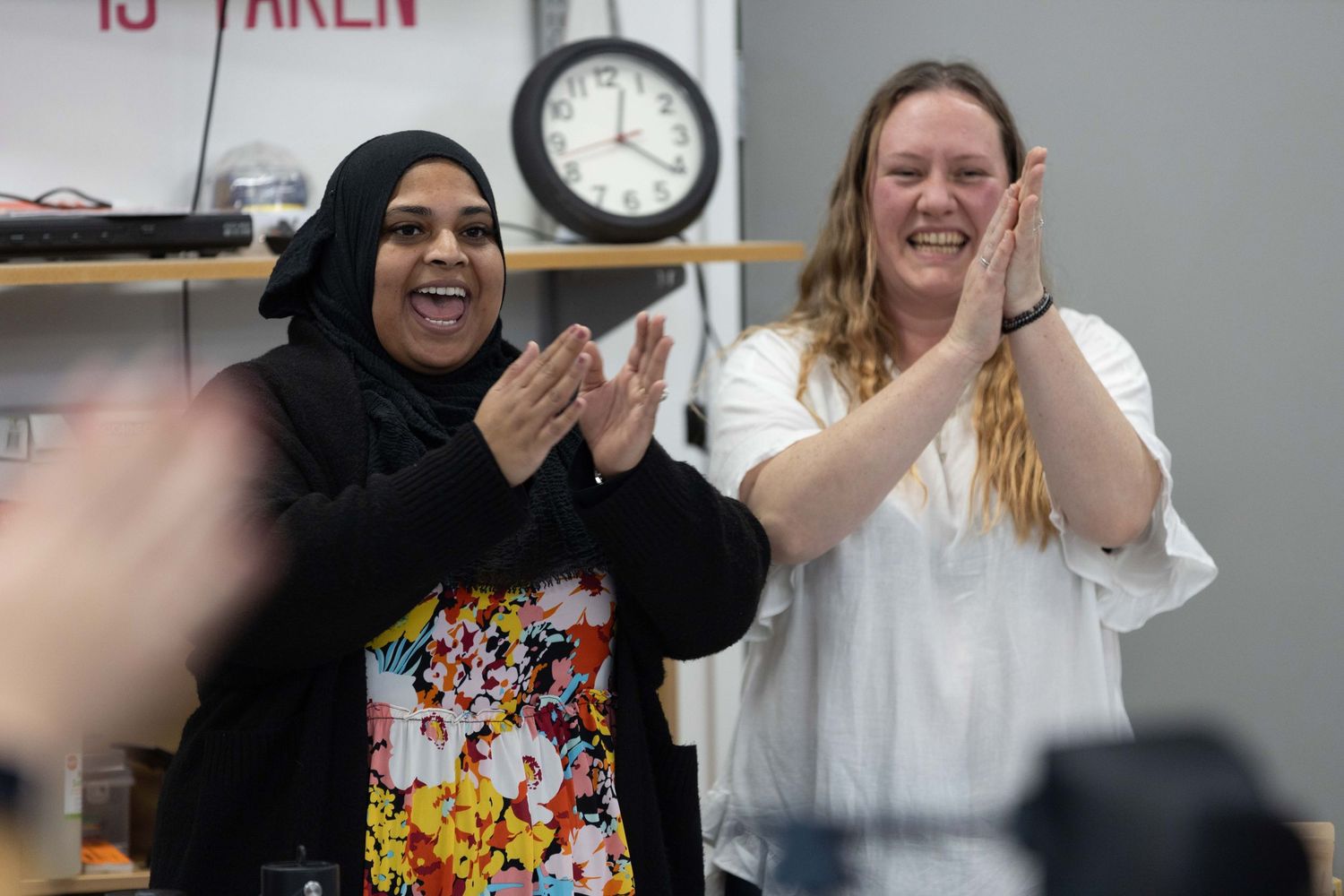 Two women clapping and smiling in a classroom, one wearing a hijab and floral top, the other in a white blouse.