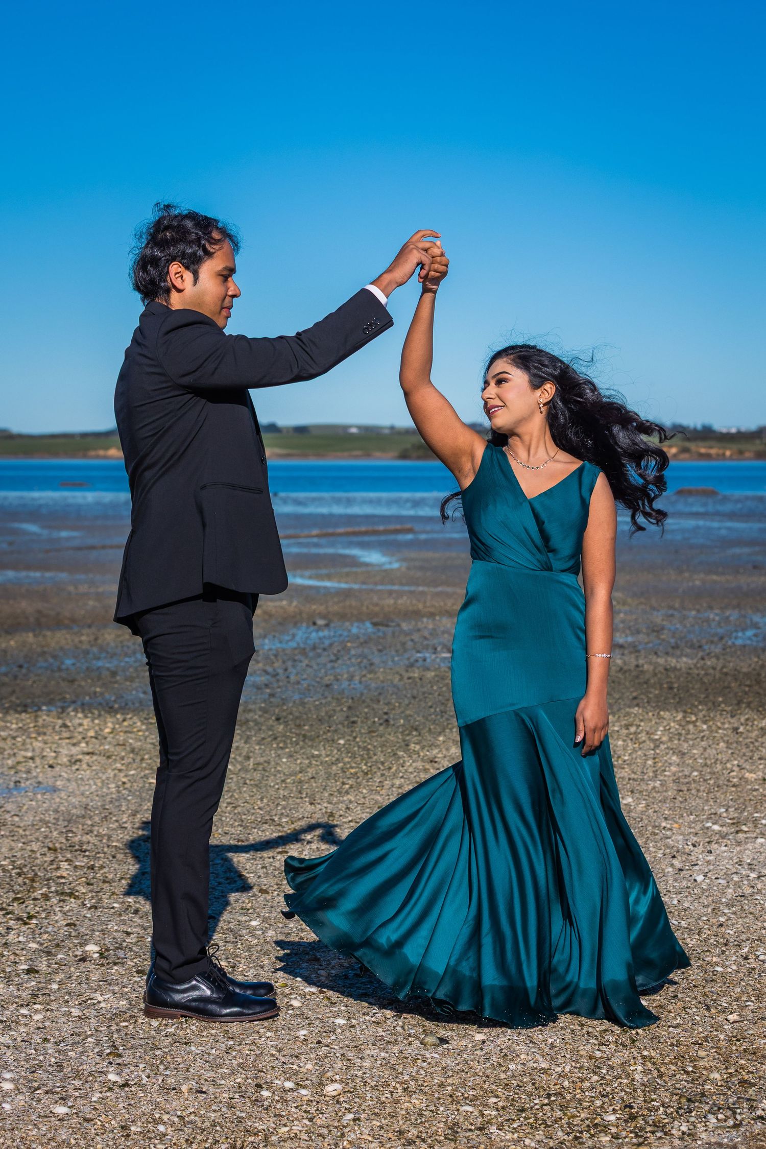 Couple dancing outdoors by a tidal flat, man in black suit spinning woman in elegant teal gown under a clear blue sky.
