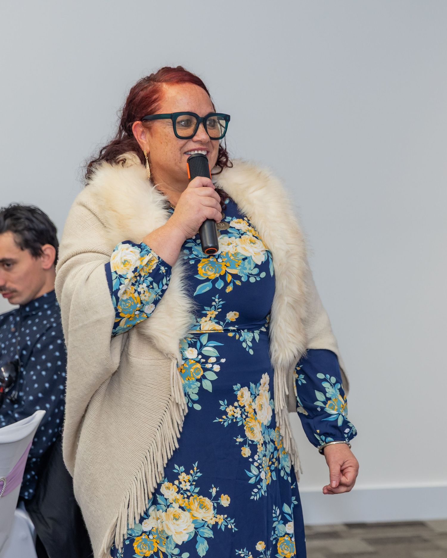 Woman in floral dress and fur shawl speaking into microphone at indoor event.