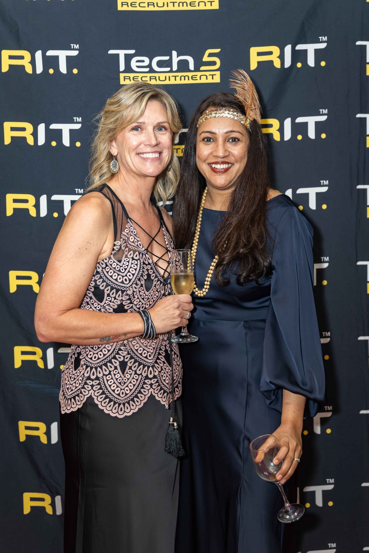Two women smiling at a Tech5 Recruitment and R.I.T. event, one holding a champagne glass, dressed in elegant attire.
