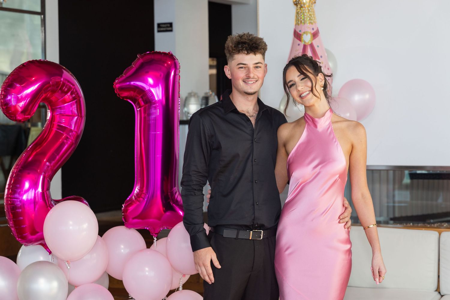 A couple poses together at a 21st birthday party with pink balloons and number 21 foil balloons in the background.