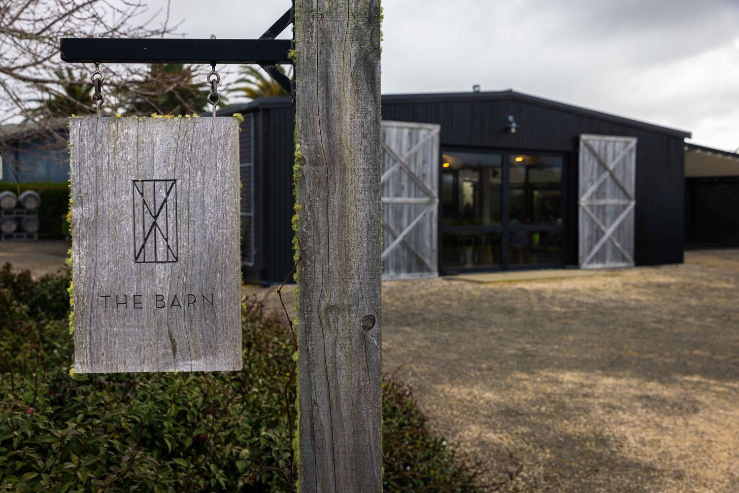 Rustic wooden sign for The Barn venue outside a dark modern barn building with sliding doors on a cloudy day.
