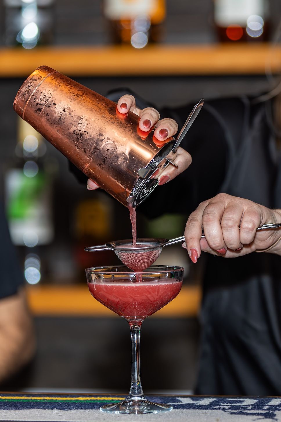 A bartender pours a vibrant red cocktail from a shaker into a coupe glass in a dimly lit bar setting.
