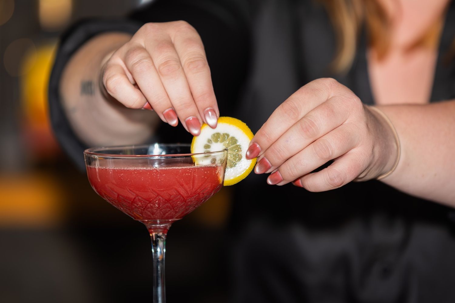 Hands garnishing a red cocktail in a martini glass with a lemon twist at a bar.