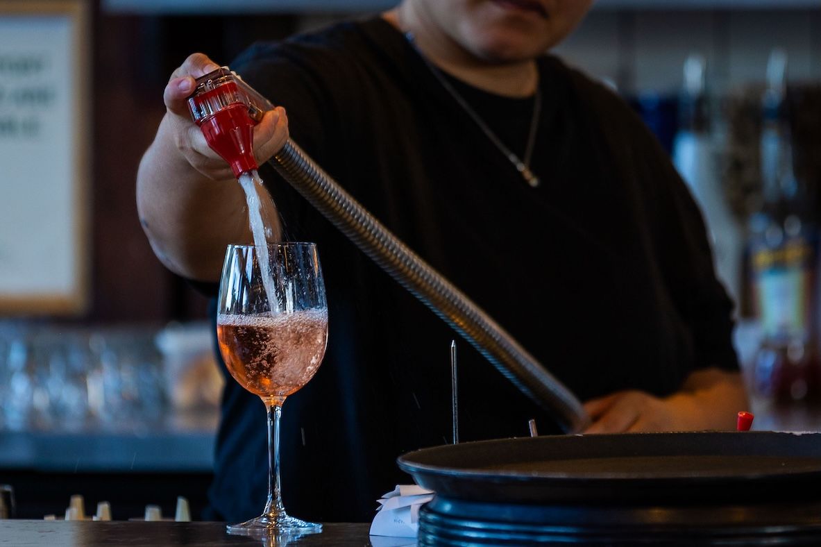 Bartender pours a vibrant orange cocktail into a wine glass at a dimly lit bar counter.
