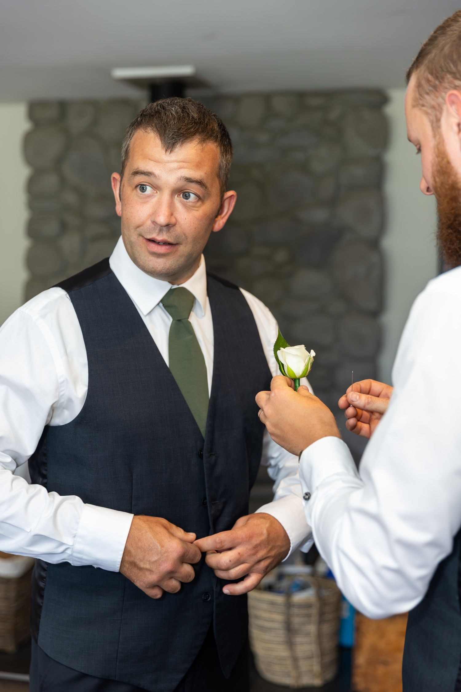 Groom in black vest and green tie gets boutonniere pinned by bearded groomsman before wedding ceremony.