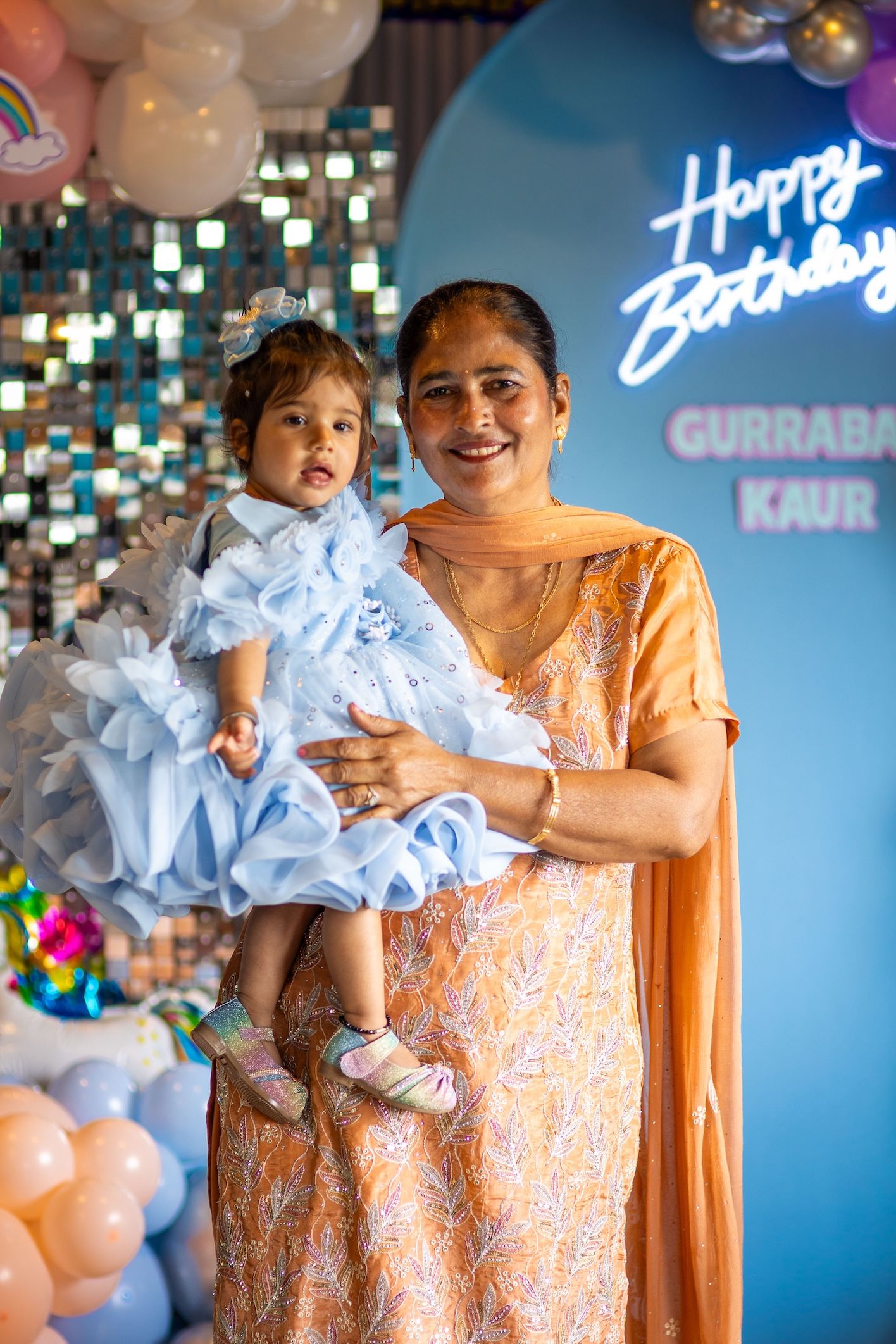 Woman in orange salwar holding a baby girl in blue ruffle dress at a Happy Birthday party celebration.