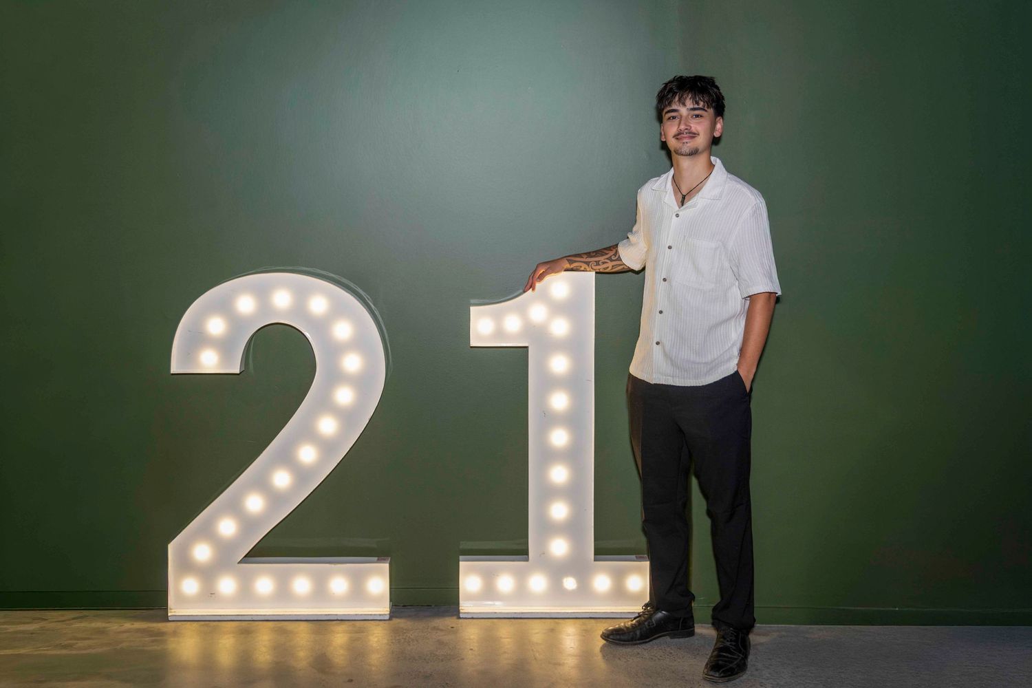 Young man posing next to illuminated number 21 marquee lights against a green wall.