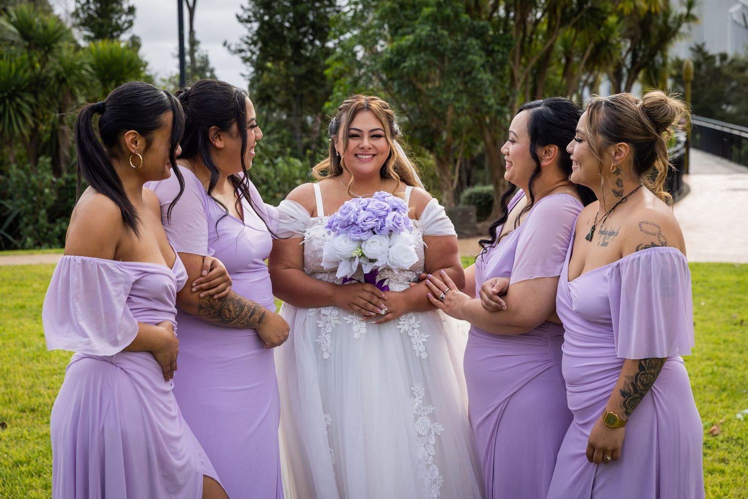 Bride in white gown holds purple bouquet surrounded by four bridesmaids in lavender off-shoulder dresses at outdoor wedding.