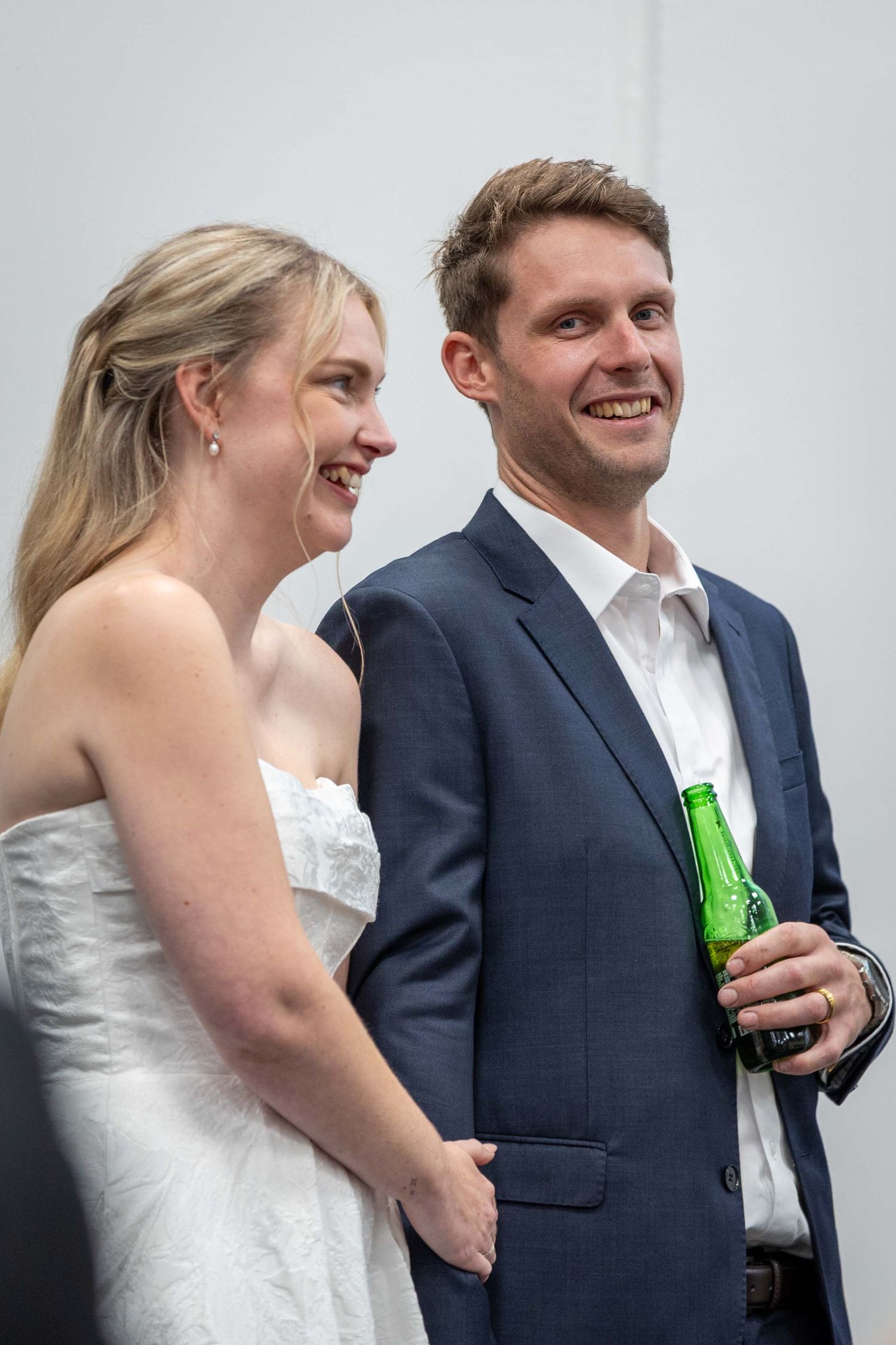 Newlyweds share candid laughing moments against white wall during reception.