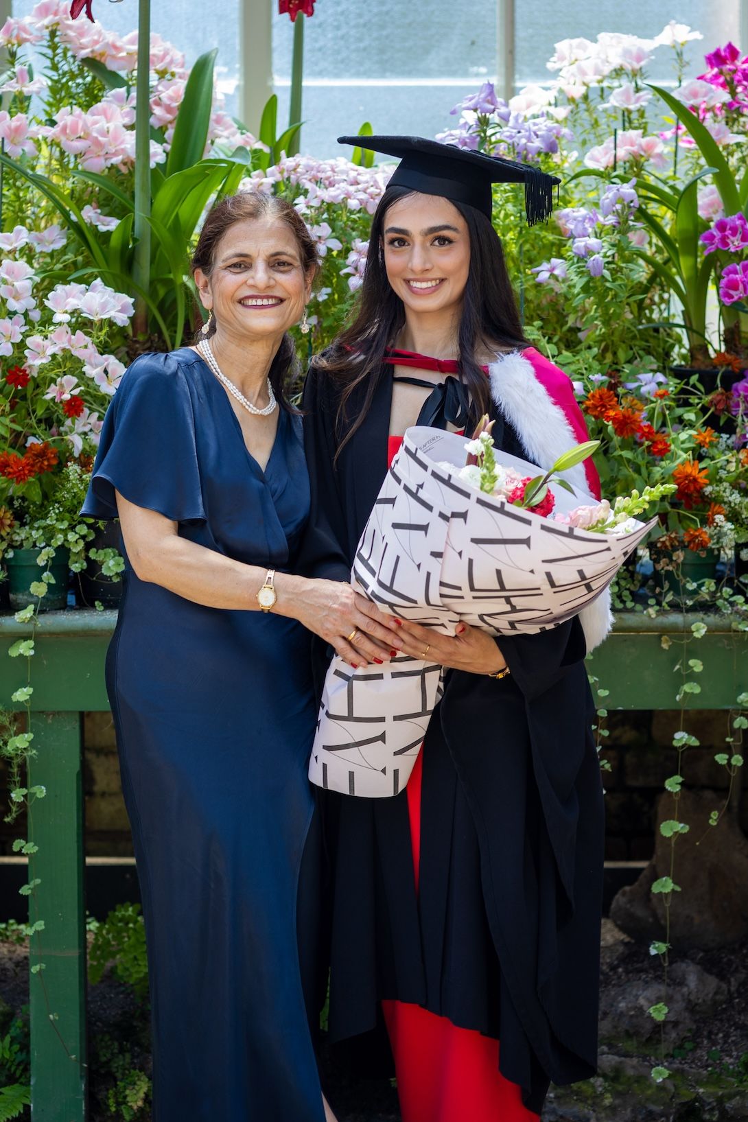Two women pose together at a graduation ceremony, one in cap and gown holding flowers, surrounded by colorful blooms.
