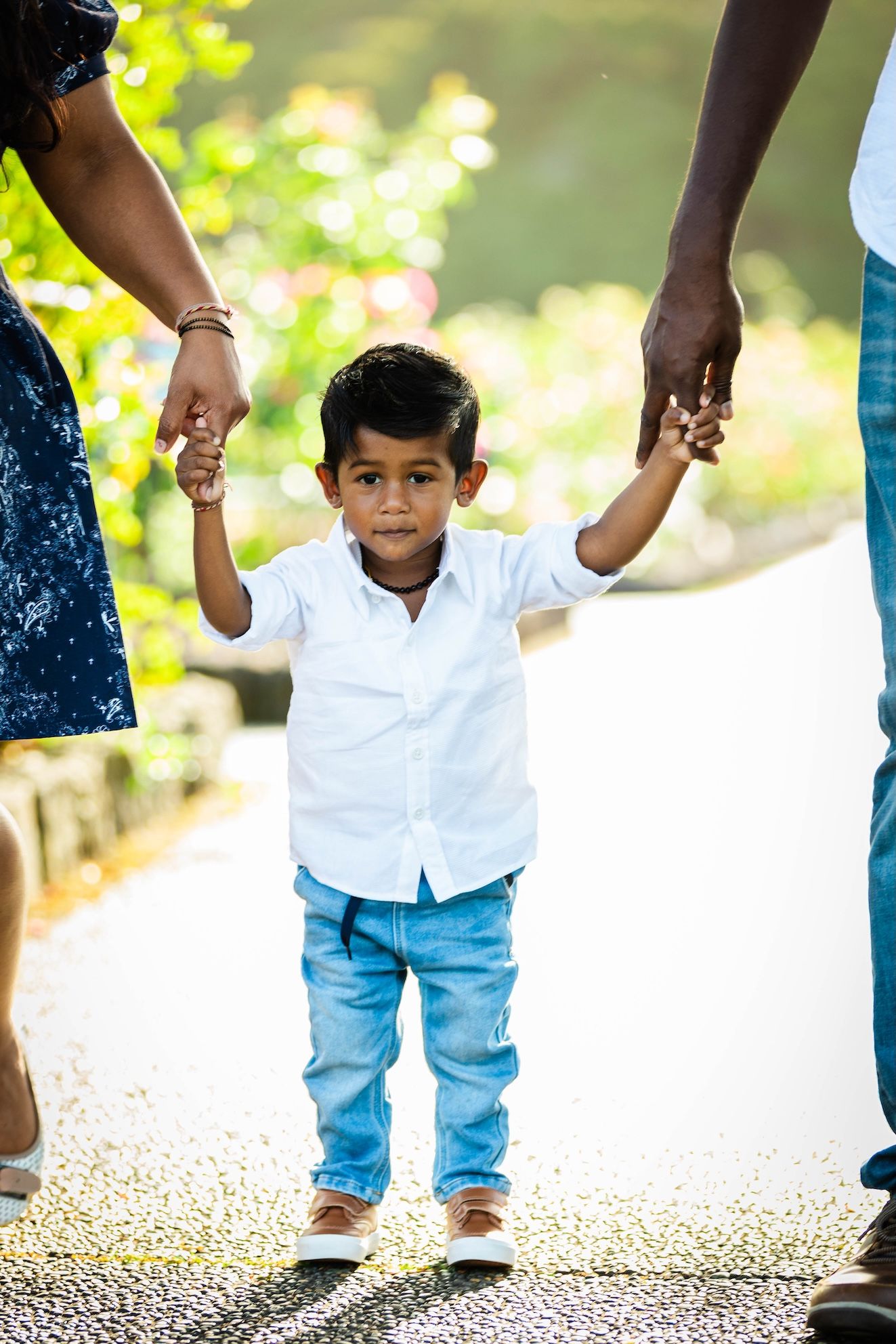 Young boy in white shirt and jeans holds hands with parents while walking outdoors in a green park.