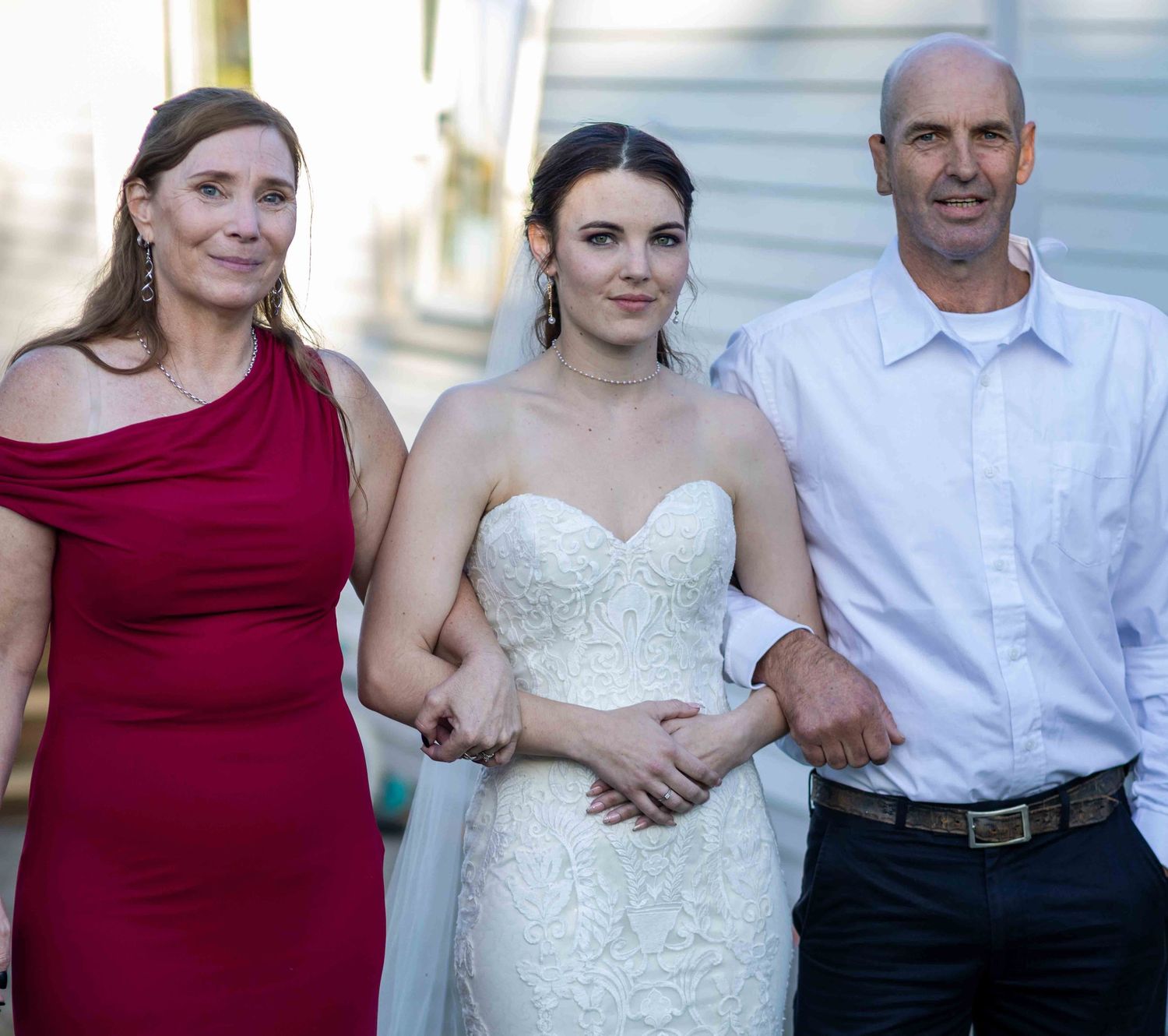 Bride in white lace gown poses with parents, mother in red dress and father in white shirt, at outdoor wedding.