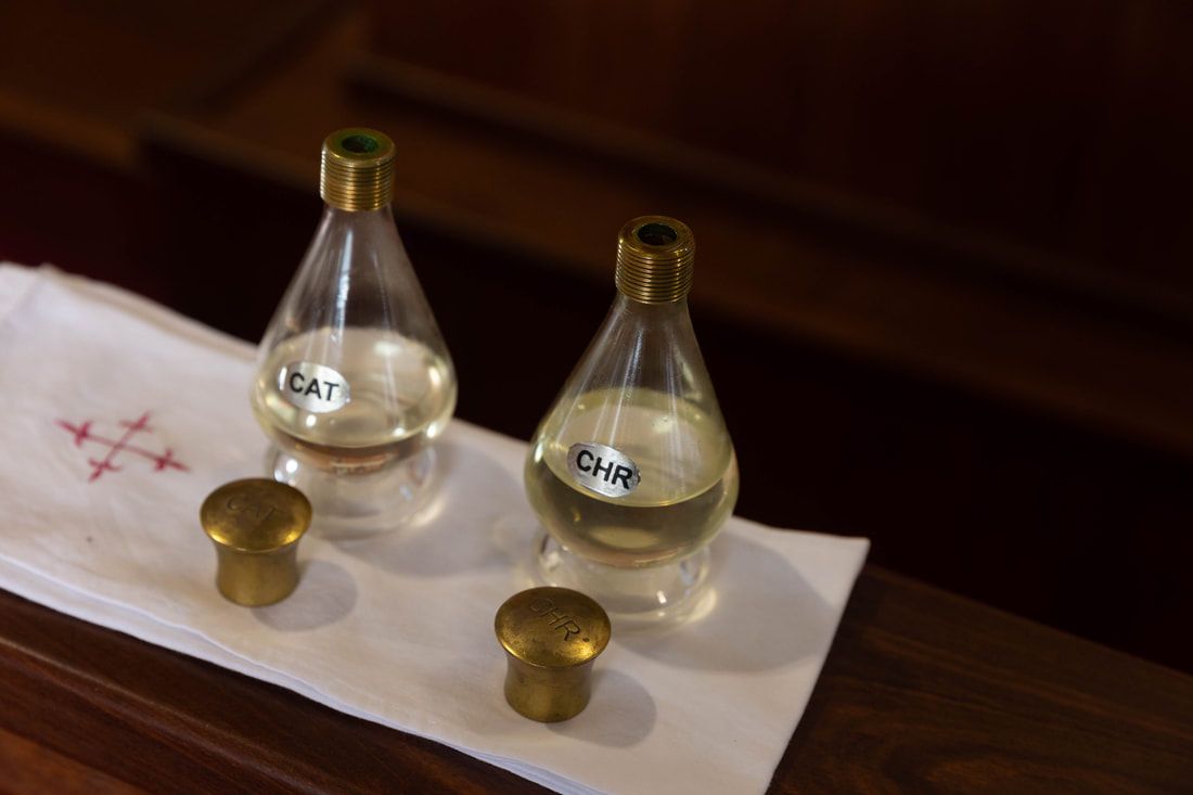 Two glass bottles labeled CAT and CHR containing holy oils on a white cloth with brass caps on a wooden surface.
