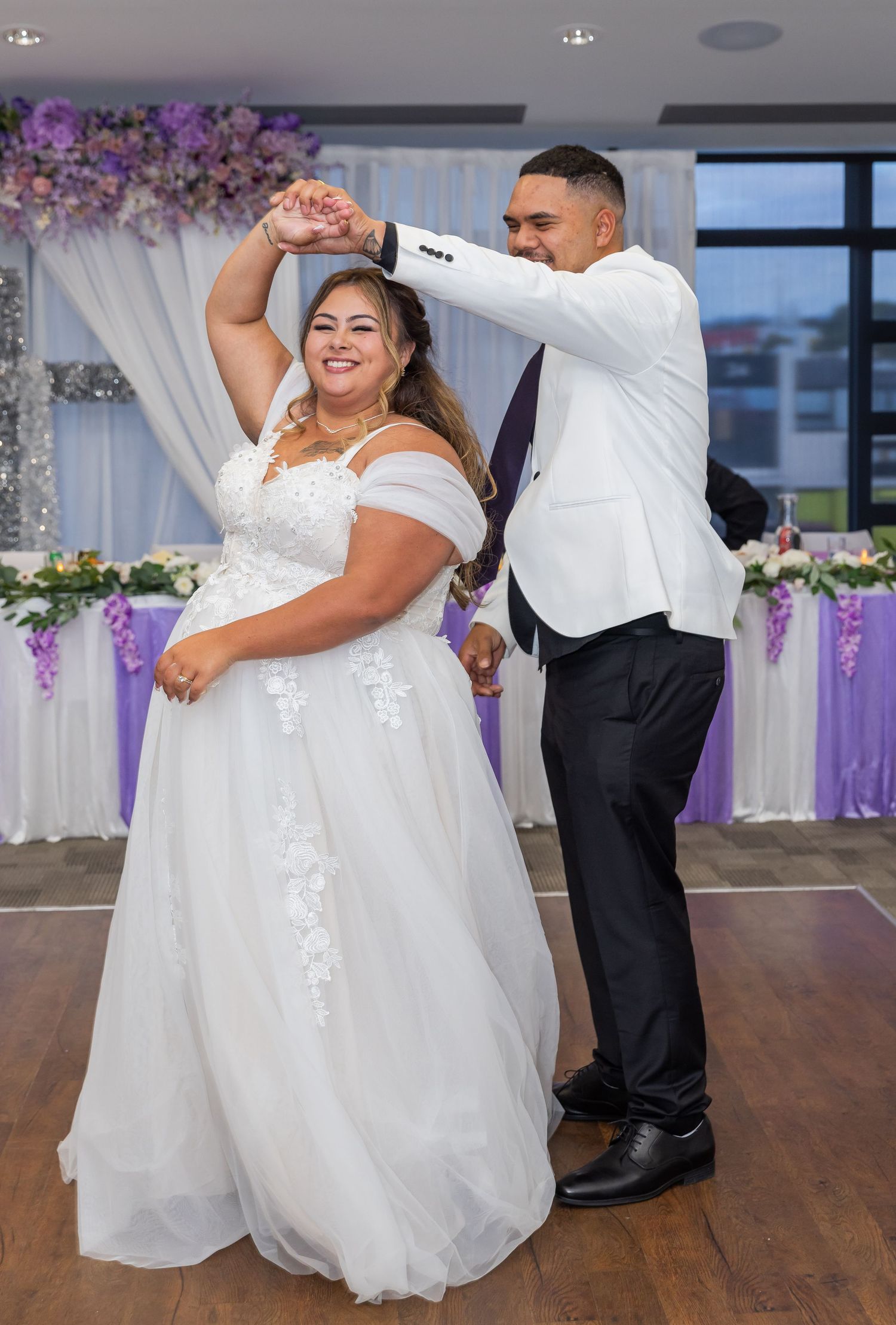 Bride and groom share their first dance at a wedding reception decorated with purple flowers and white draping.