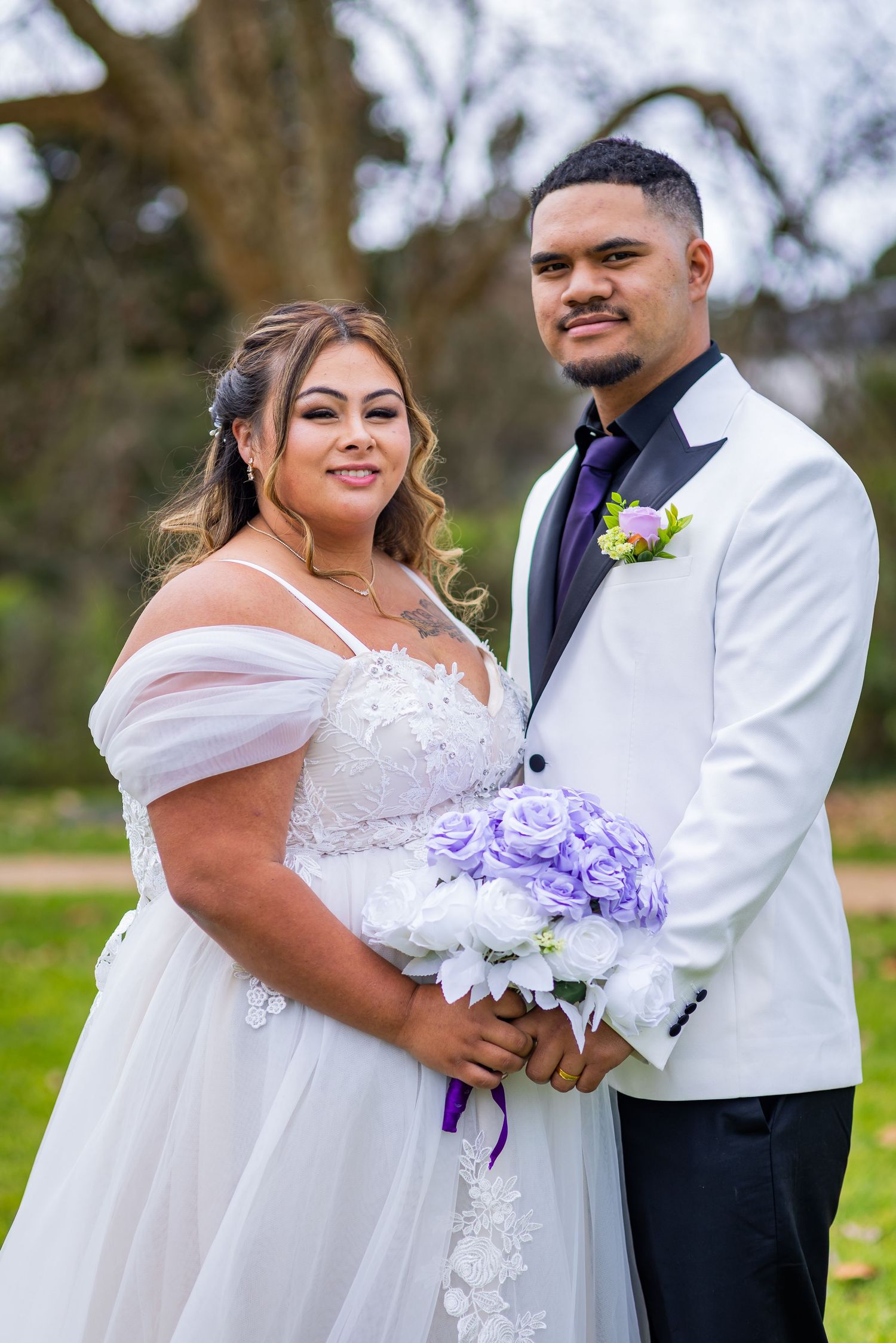 Bride in white lace off-shoulder gown holds purple bouquet beside groom in white tuxedo outdoors.