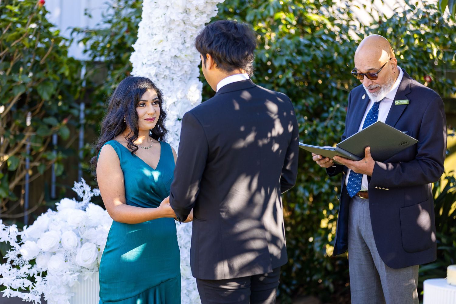 Couple exchanging wedding vows outdoors with officiant reading from book, white floral arch backdrop.