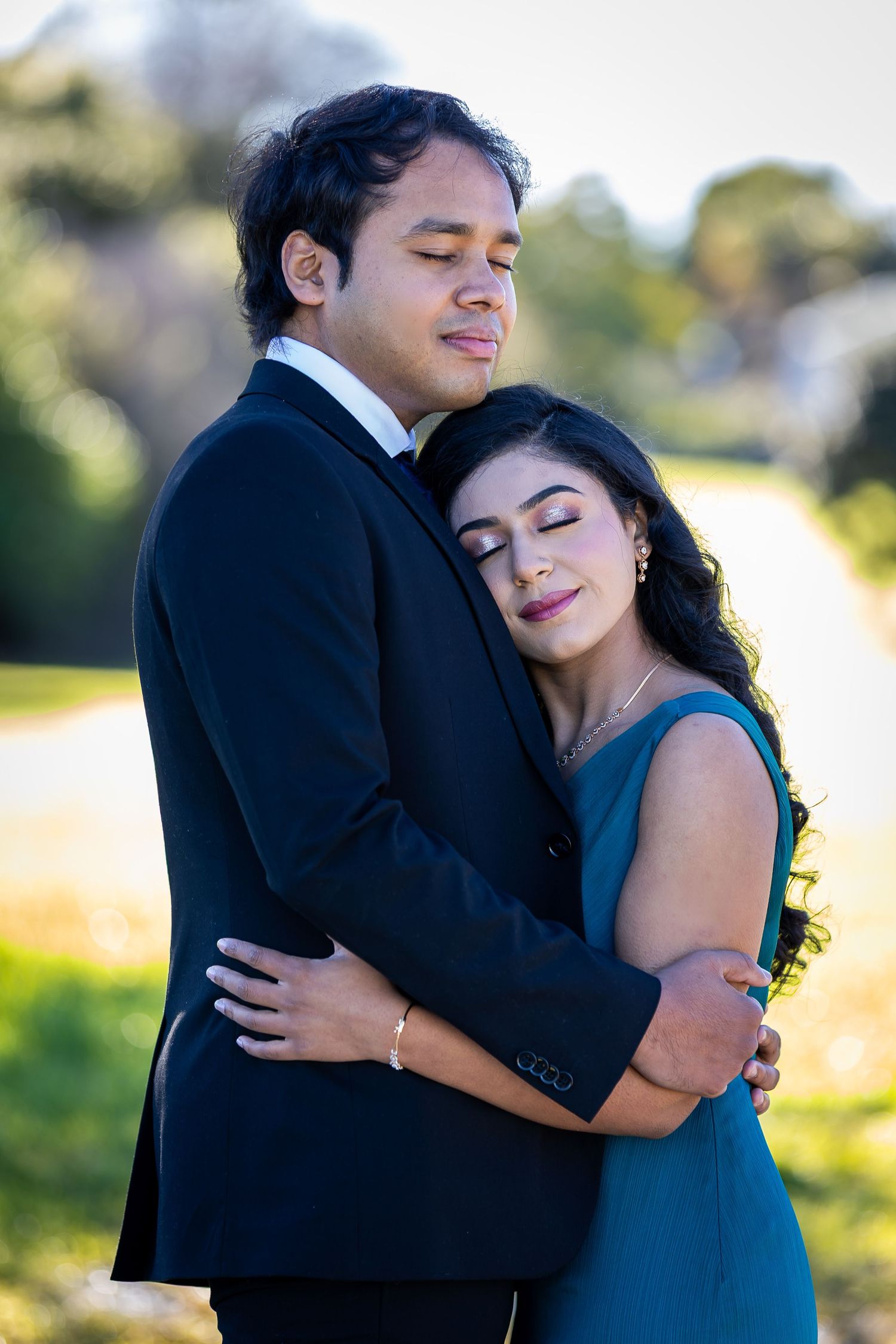 Couple in formal attire embracing outdoors, woman in teal dress resting head on man's chest in black suit.