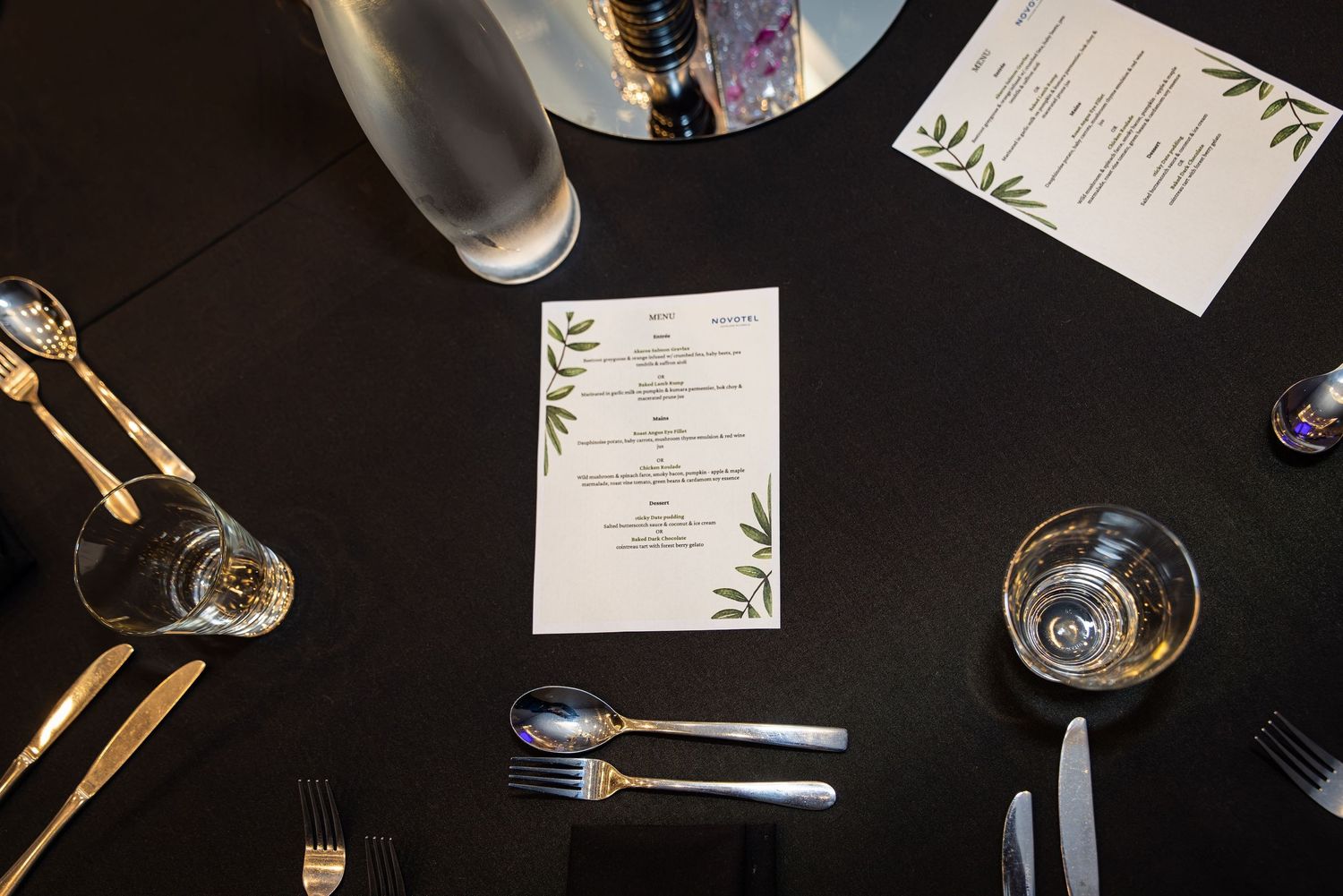 Elegant black table setting with gold cutlery, glassware, and a decorative menu card with green leaf accents.