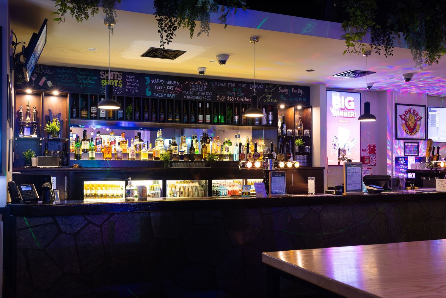Wide panoramic view of an empty modern bar interior with purple ambient lighting.