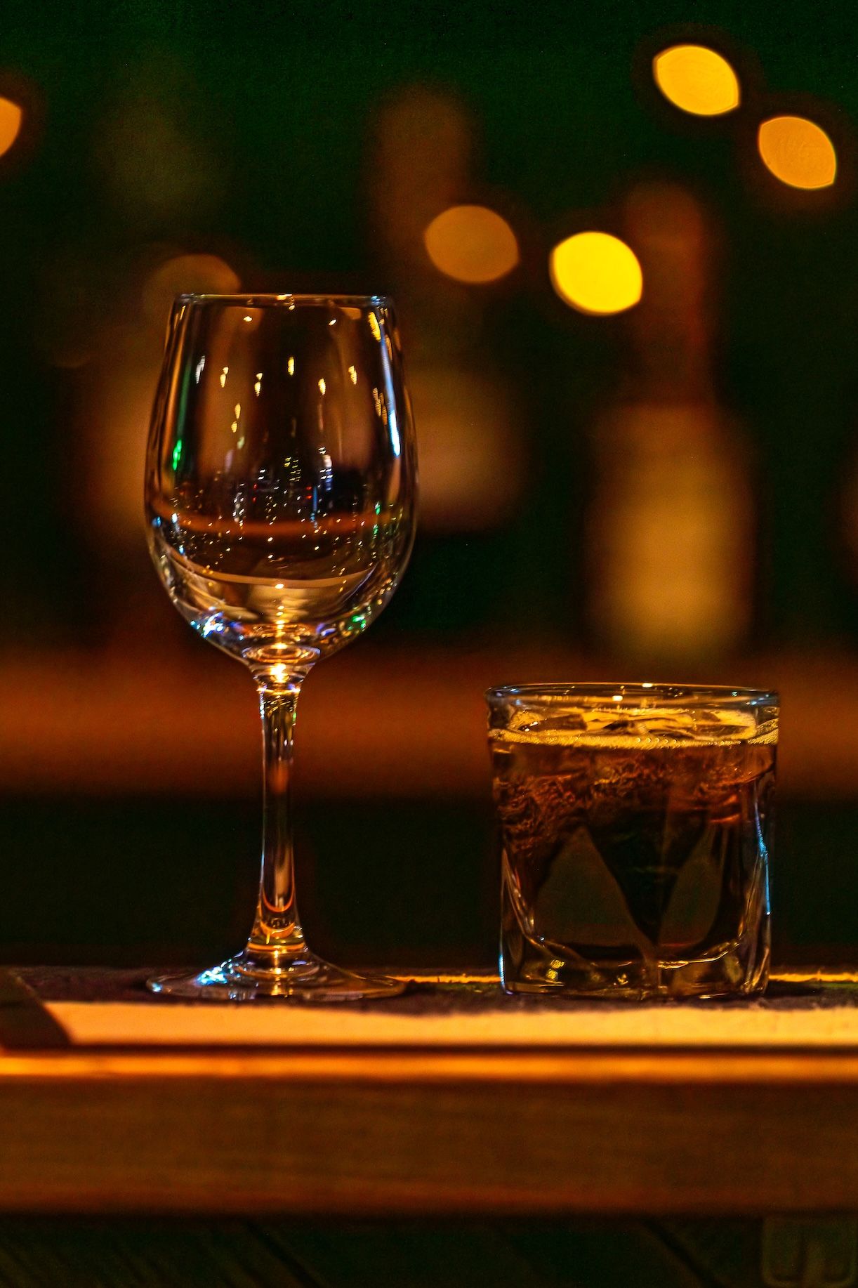 Wine glasses sit on a dark wooden bar counter with warm ambient lighting and blurred bokeh in the background.