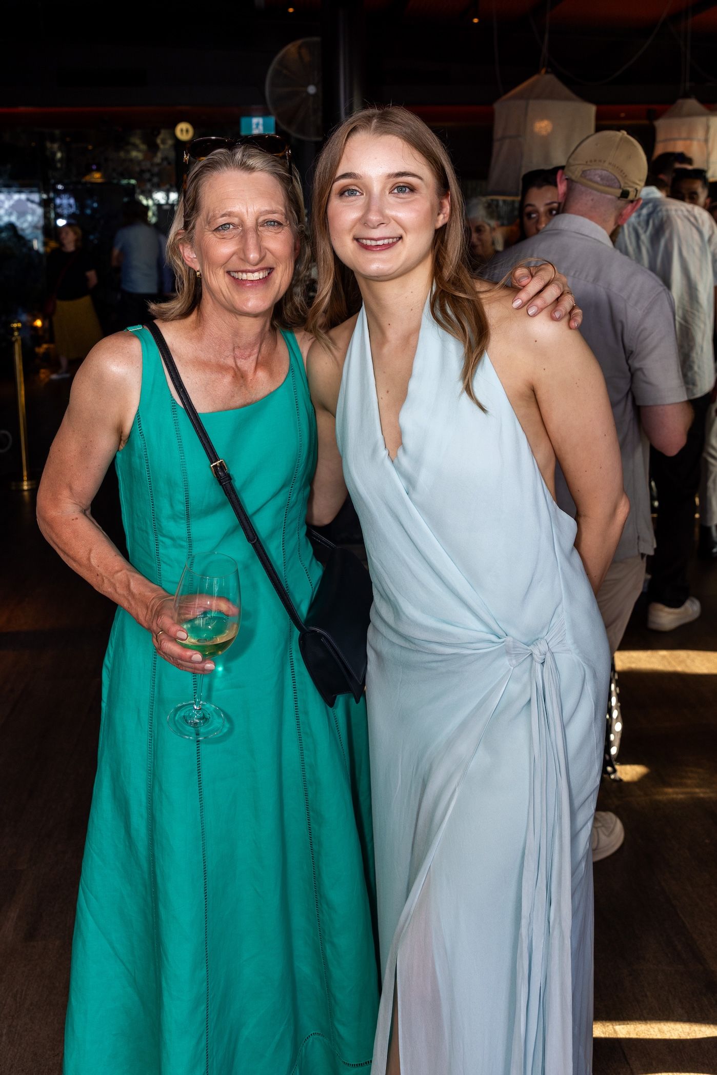 Two women in elegant dresses smile together at a formal event, one in teal and one in light blue.