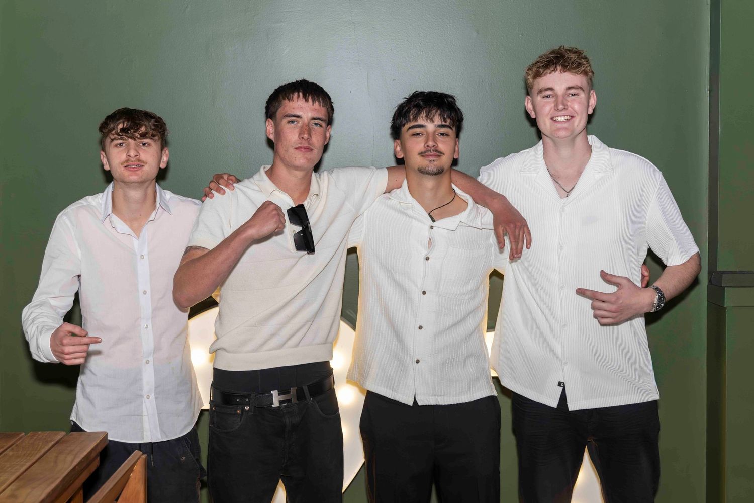 Four young men in white shirts pose together smiling against a green wall.