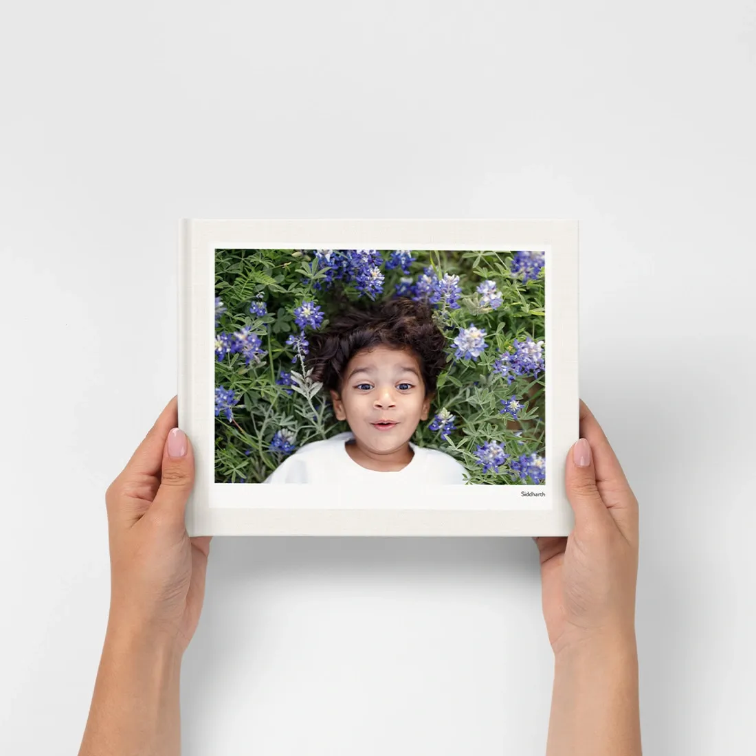 Hands holding a framed photo of a child lying in a field of purple wildflowers, smiling up at the camera.