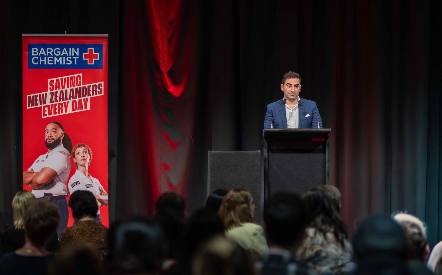 Speaker at podium presenting to audience with Bargain Chemist banner visible.