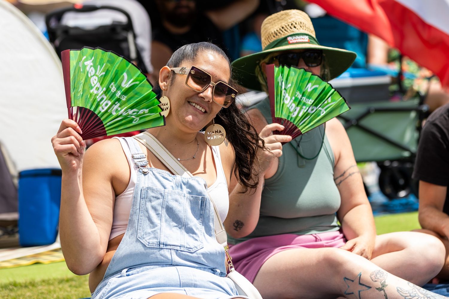 Festival attendees sitting on grass with green promotional fans on sunny day.
