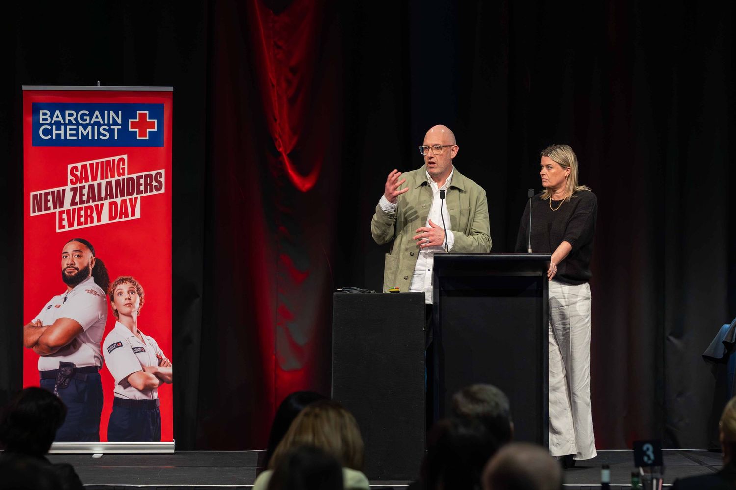 Corporate presentation with speakers on stage featuring Bargain Chemist branding and red lighting.