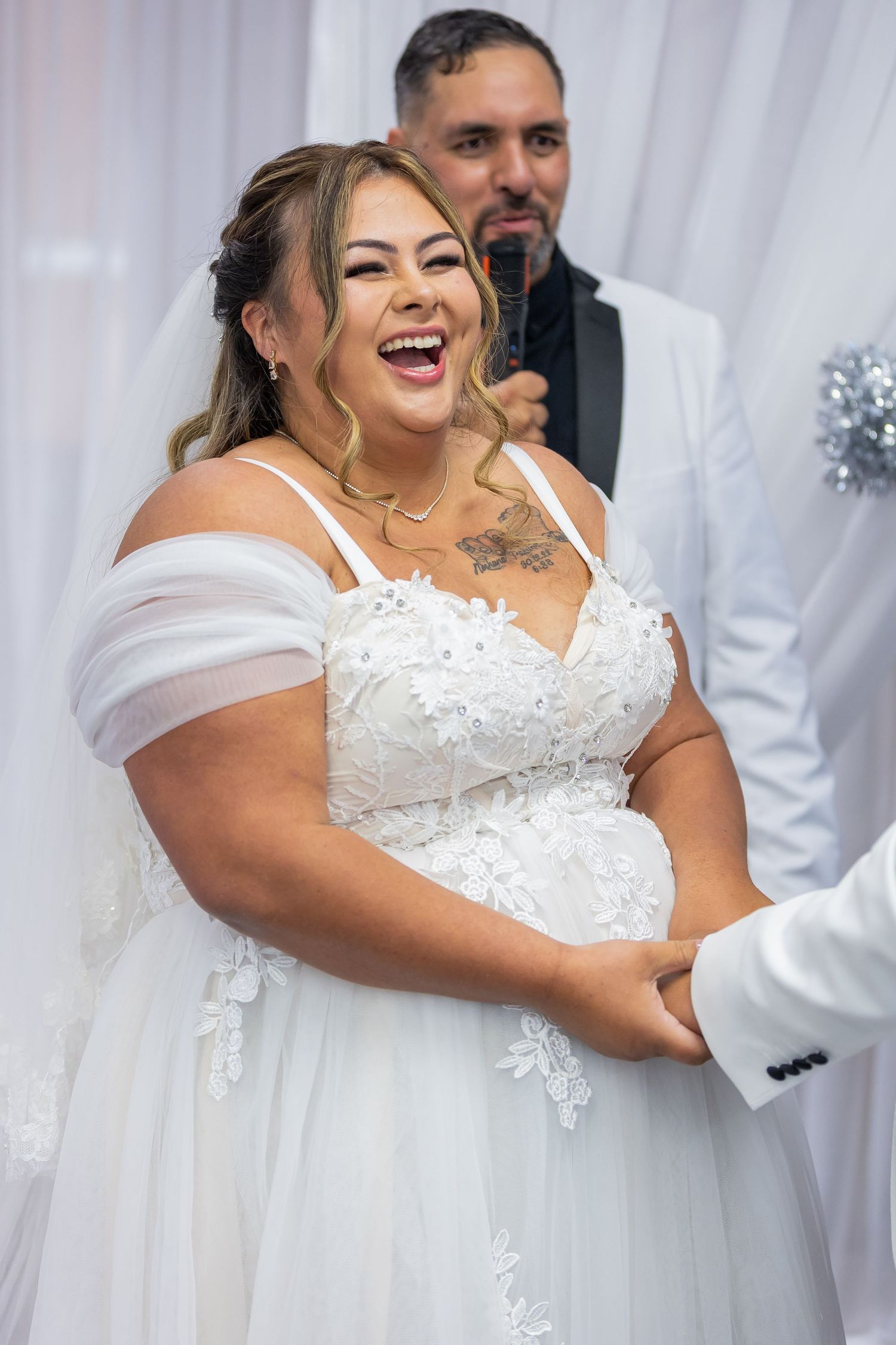 Laughing bride in white off-shoulder lace gown holds hands with groom in white suit at wedding ceremony.