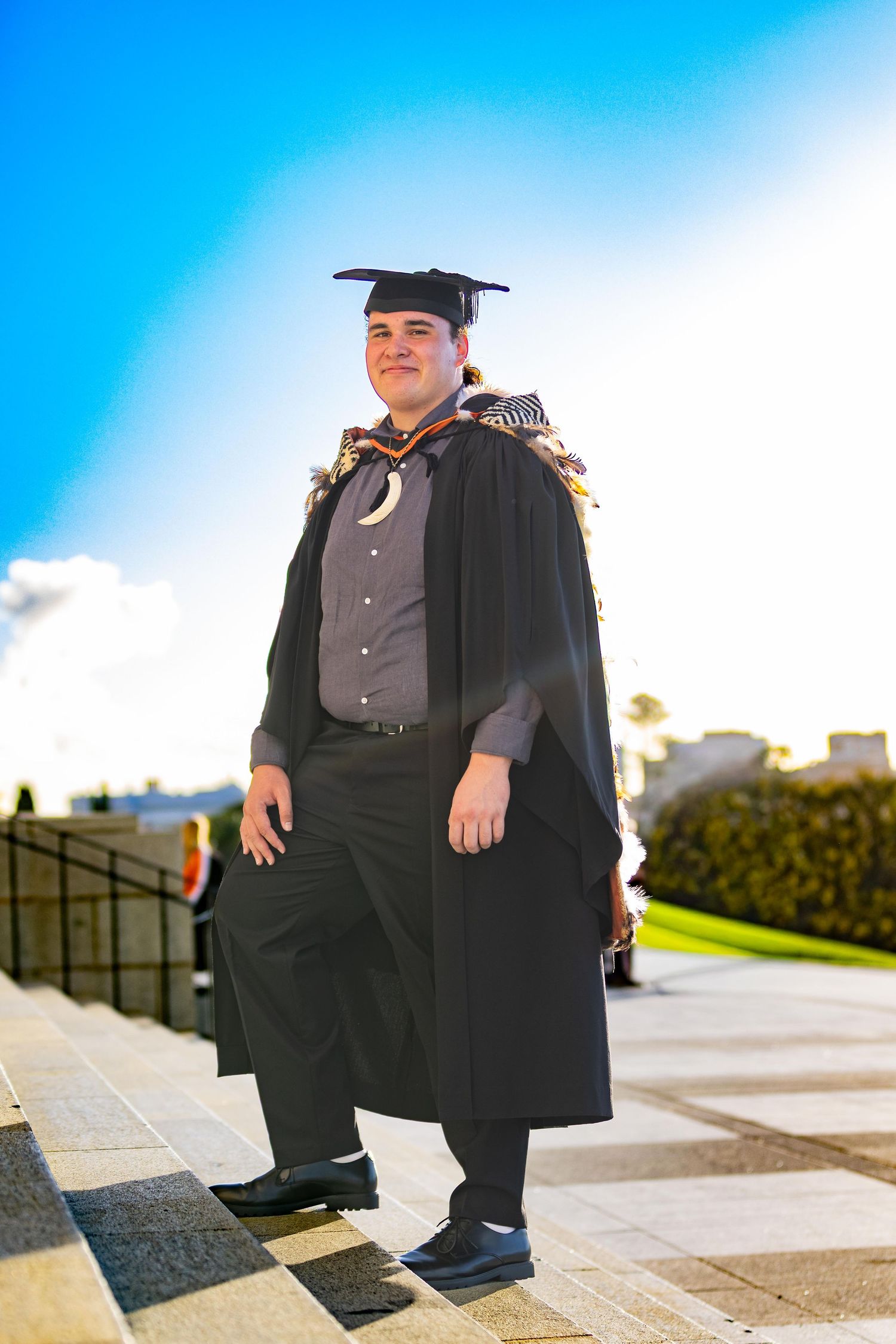 Graduate in black cap and gown smiles outdoors on sunny day, wearing academic regalia with decorative hood.
